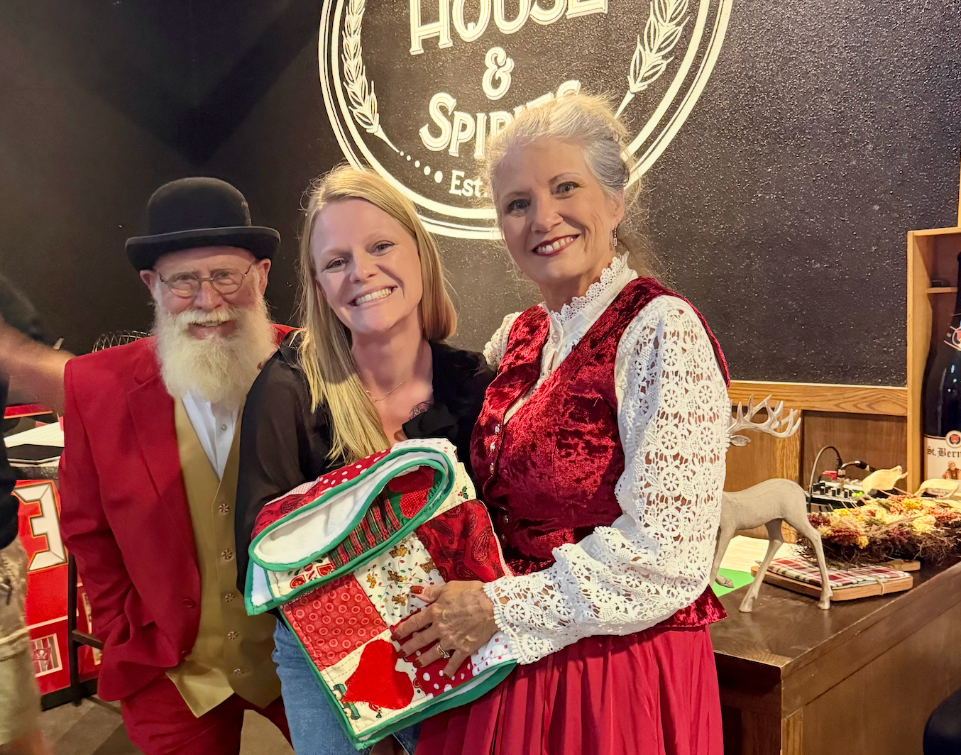 Three people in festive attire pose together holding a holiday gift bag at House and Spirits establishment.