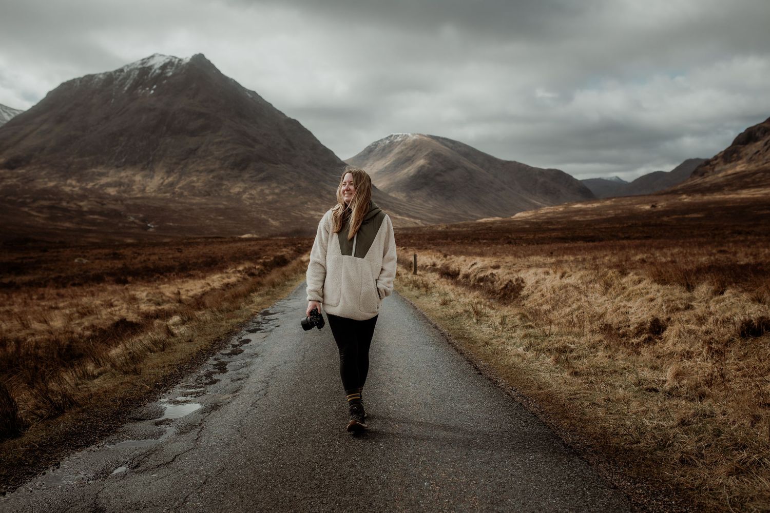 Woman walking on a narrow road through a dramatic Scottish Highland landscape with mountains and overcast skies.