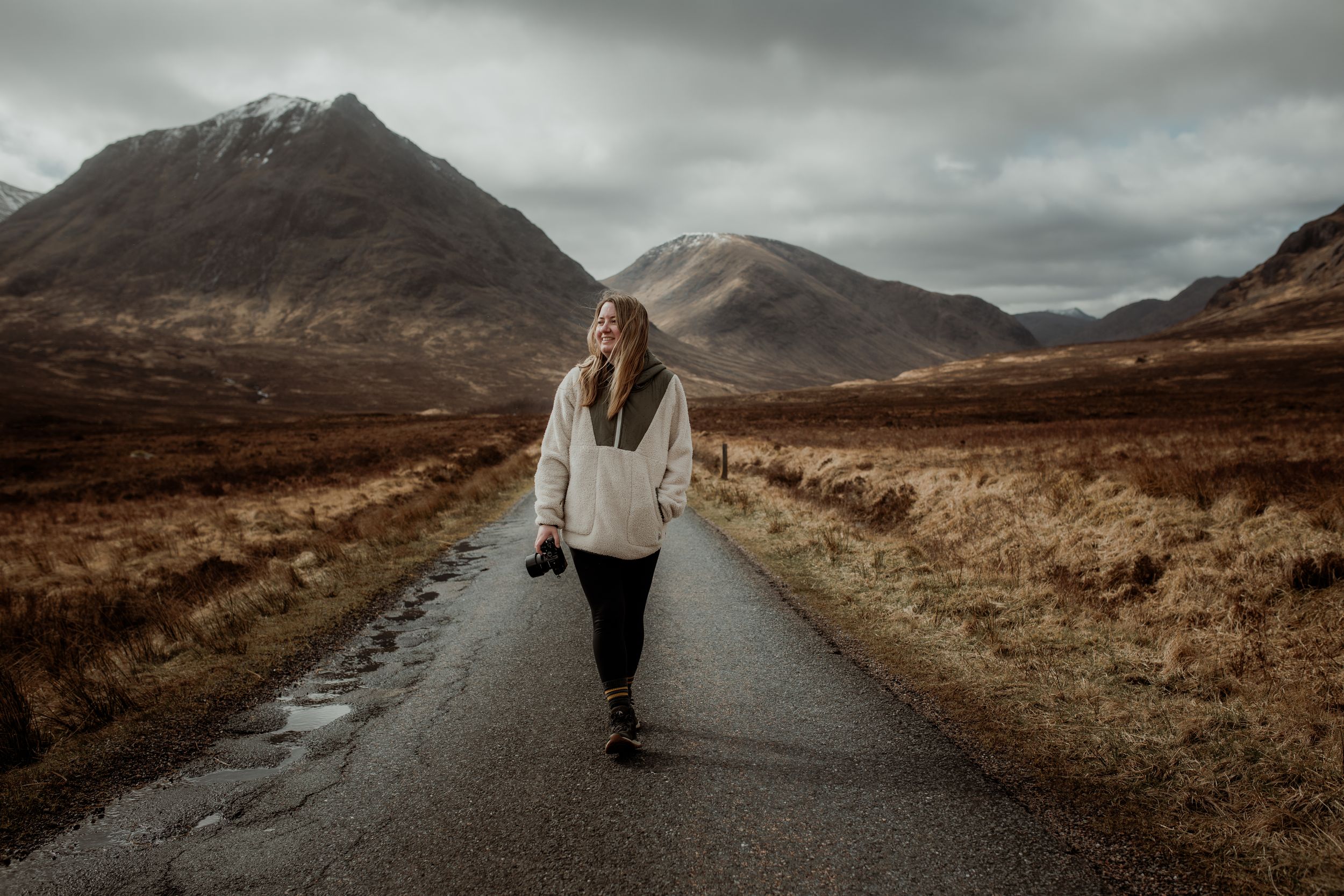 Woman walking on a narrow road through a dramatic Scottish Highland landscape with mountains and overcast skies.
