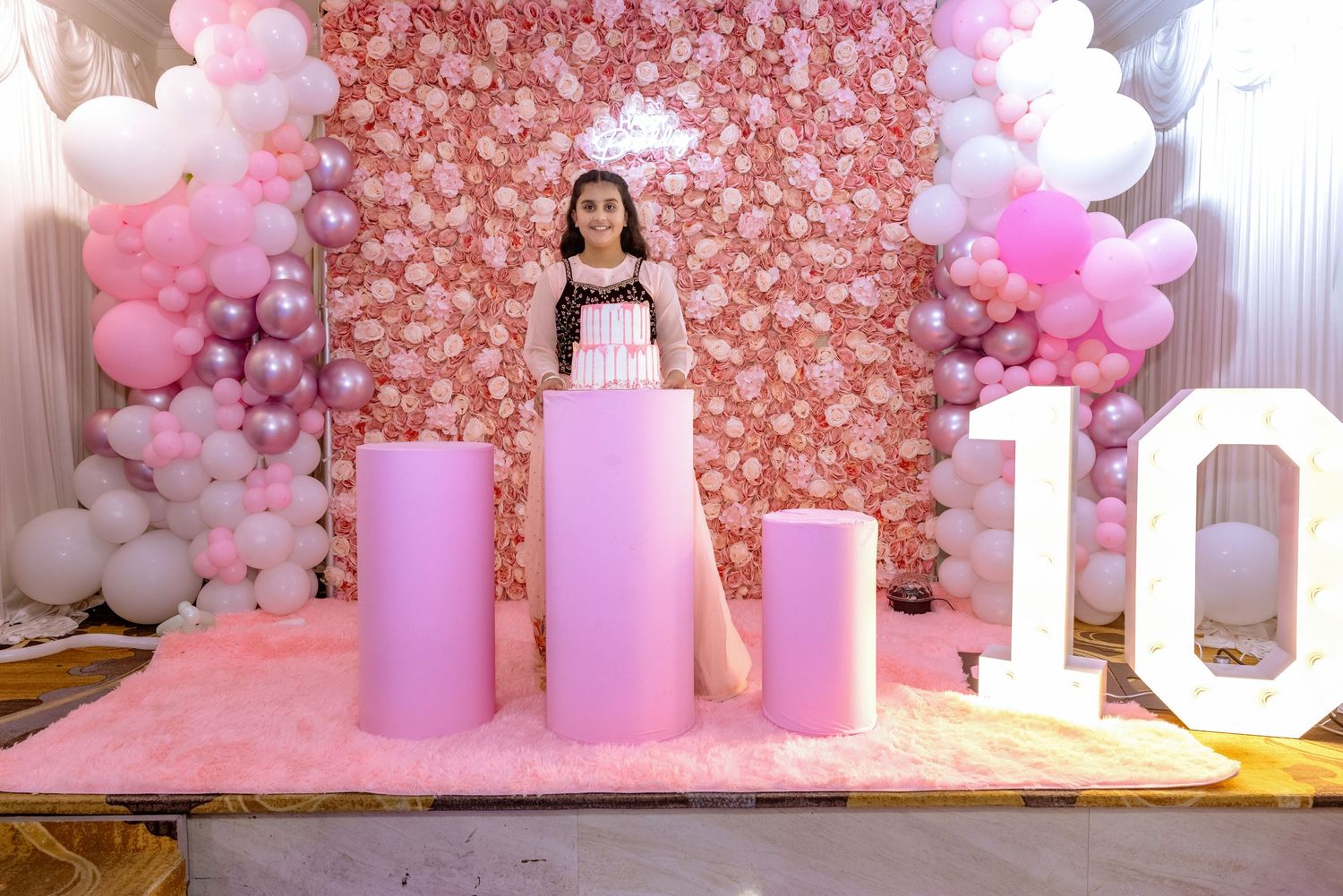 Girl celebrates 10th birthday with pink balloon arch, floral wall backdrop, and decorative pedestals at a party.