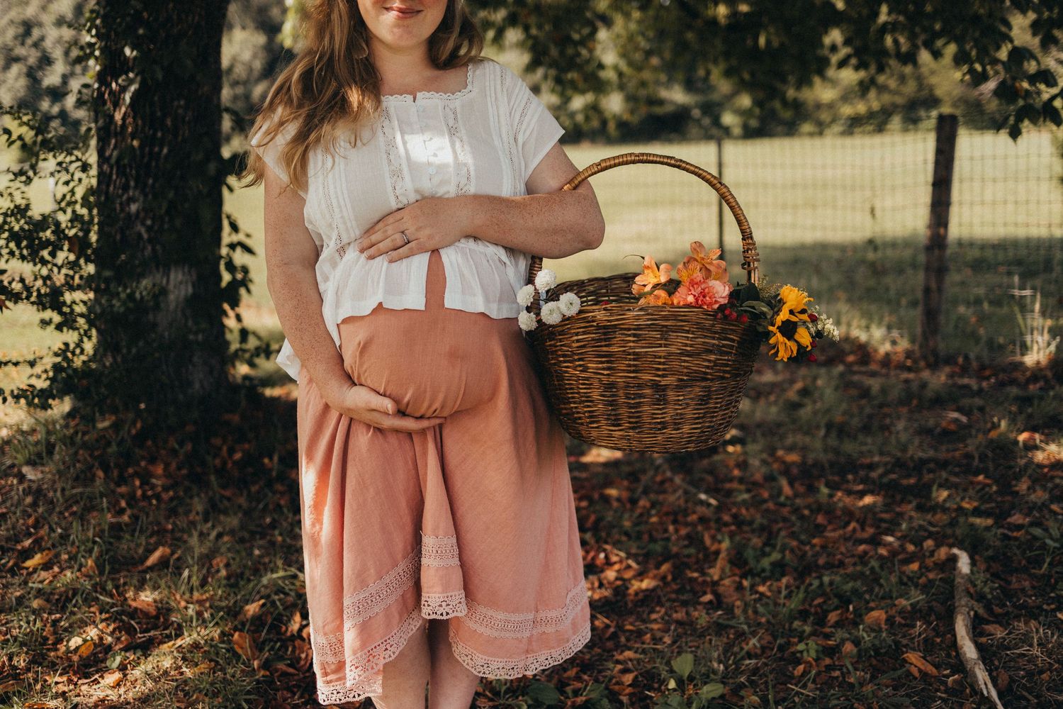 Pregnant woman in white top and peach skirt holding a wicker basket with flowers outdoors in a sunlit garden in Knoxville, TN.