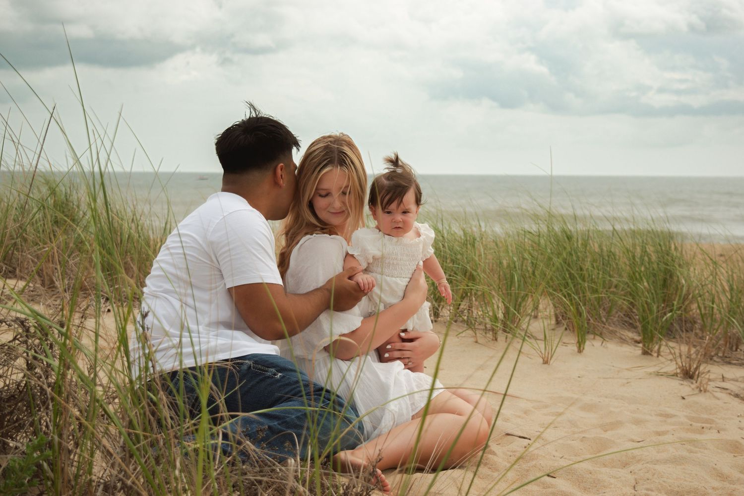 A young family sits on sandy beach dunes, parents admiring their baby girl with ocean waves in the background.
