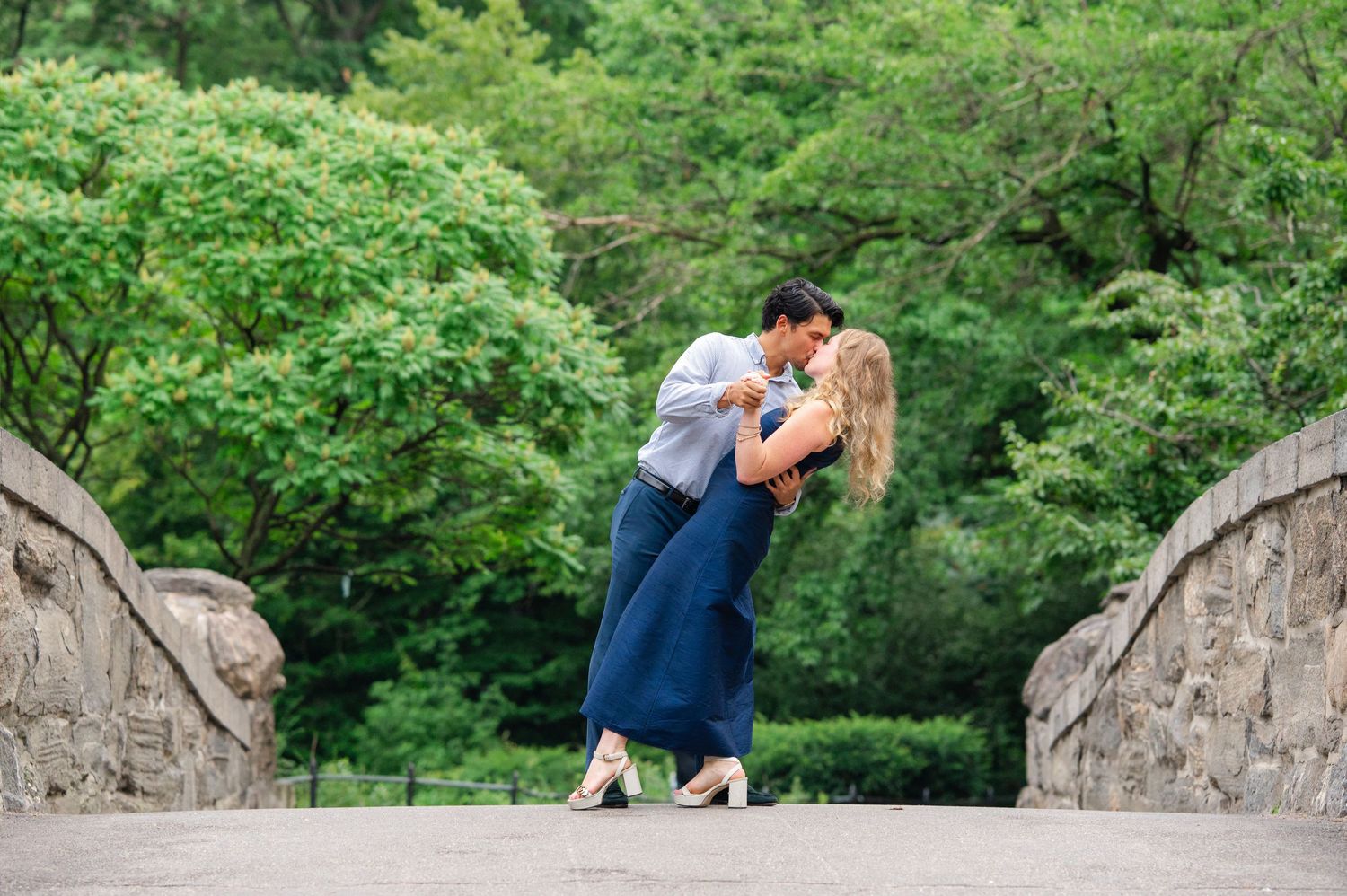 Couple sharing a romantic dip kiss on a stone bridge surrounded by lush green trees in Central Park.
