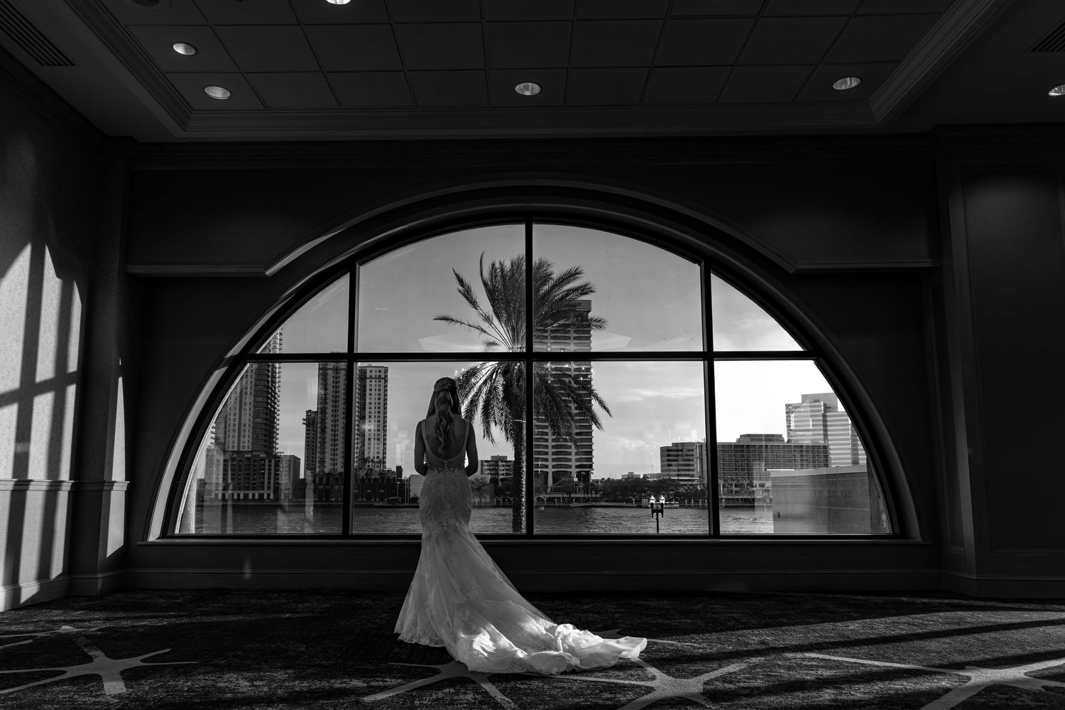 Bride in wedding gown stands before arched window with palm tree and city skyline in elegant black and white photo.