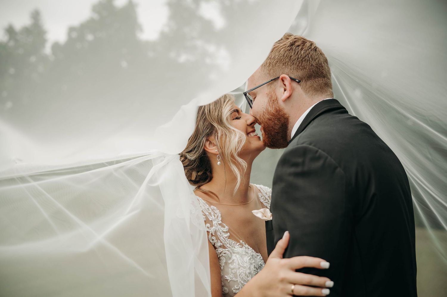 Bride and groom sharing an intimate moment under a flowing white veil, nearly kissing in soft natural light.