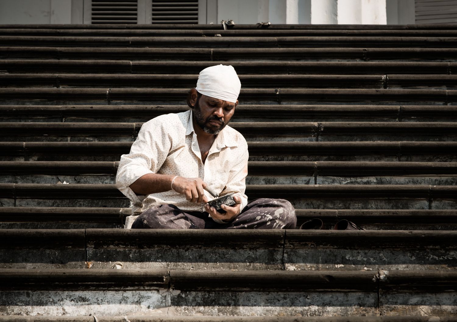 Indian man in white clothes sitting on stone steps examining something in his hands.