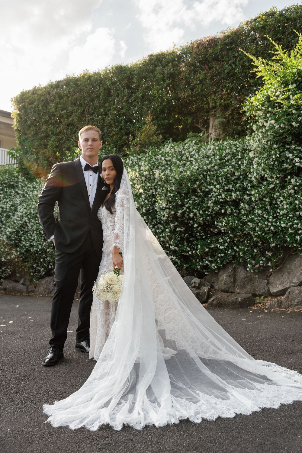 A romantic wedding couple poses together outdoors against a lush green hedge backdrop with a flowing lace veil trailing behind.