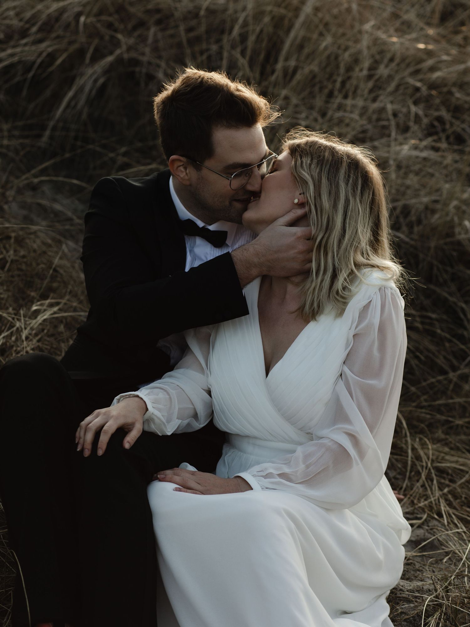 A romantic outdoor portrait of a couple in formal attire embracing in tall grass at sunset.