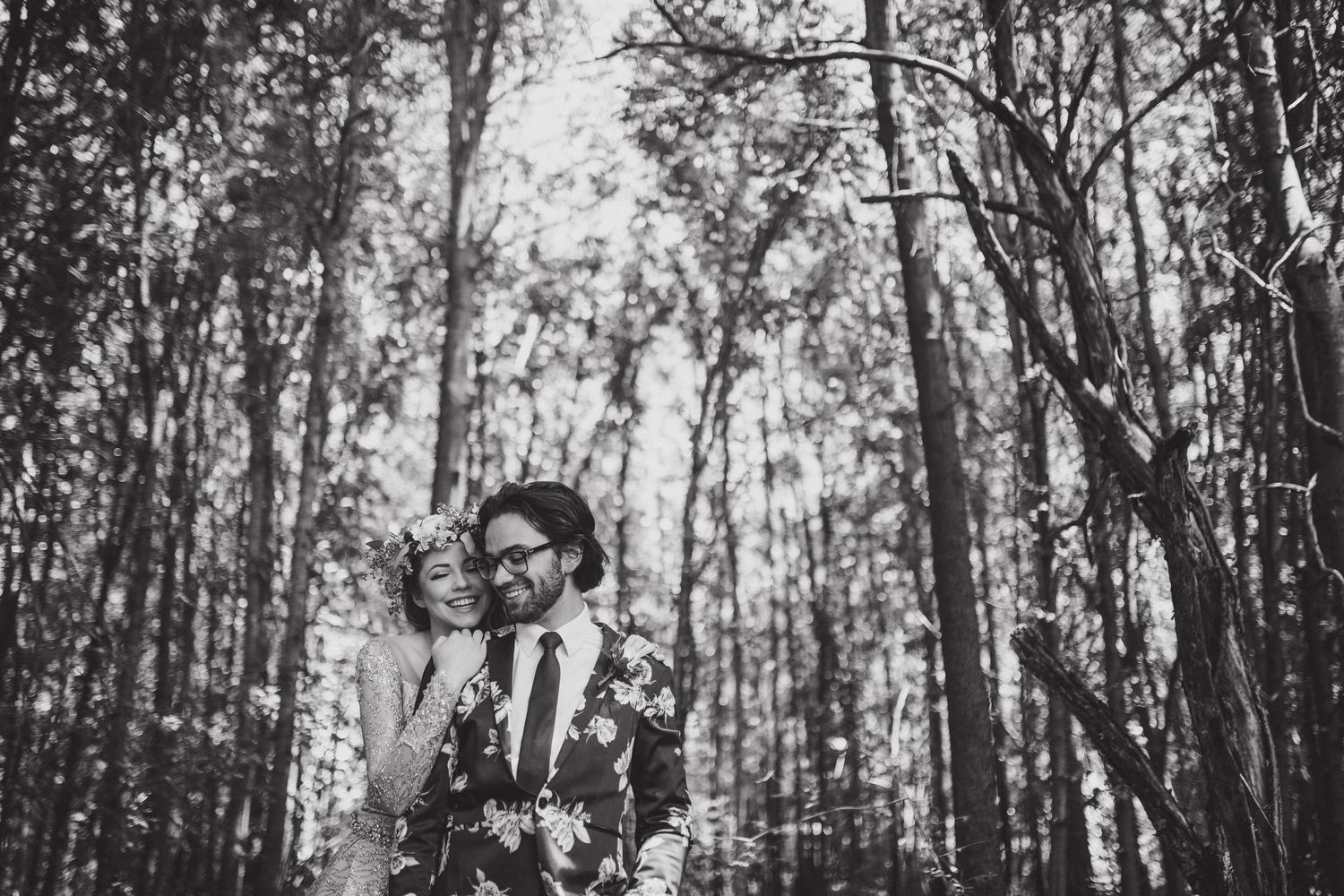 A black and white portrait of a couple standing among tall trees in a forest.