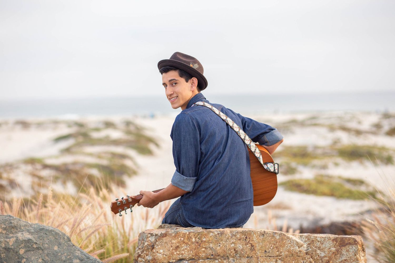 A musician in a blue shirt sits with a guitar on rocks overlooking a scenic landscape.