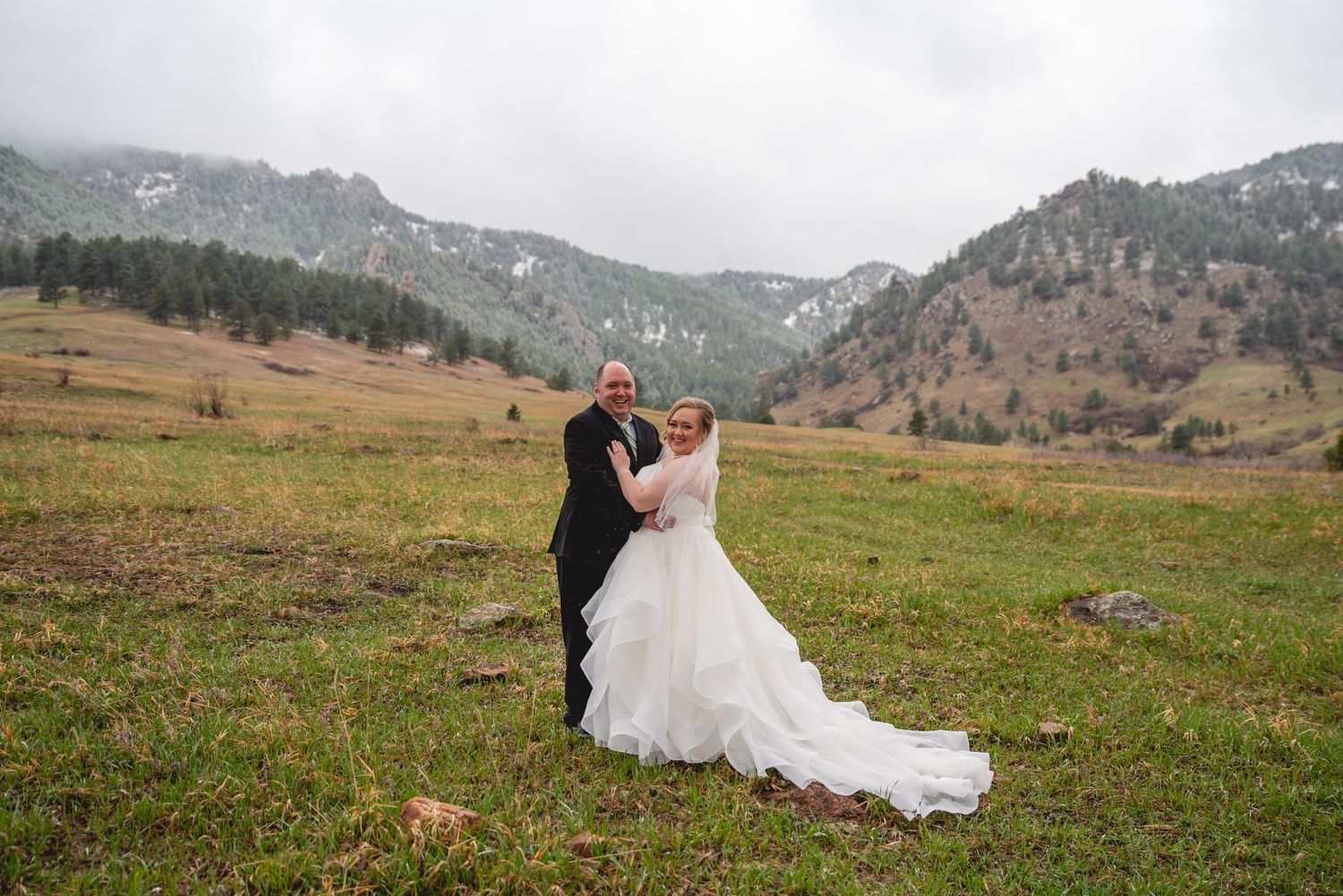 A couple in wedding attire embraces on a grassy hillside with snow-capped mountains in the background.