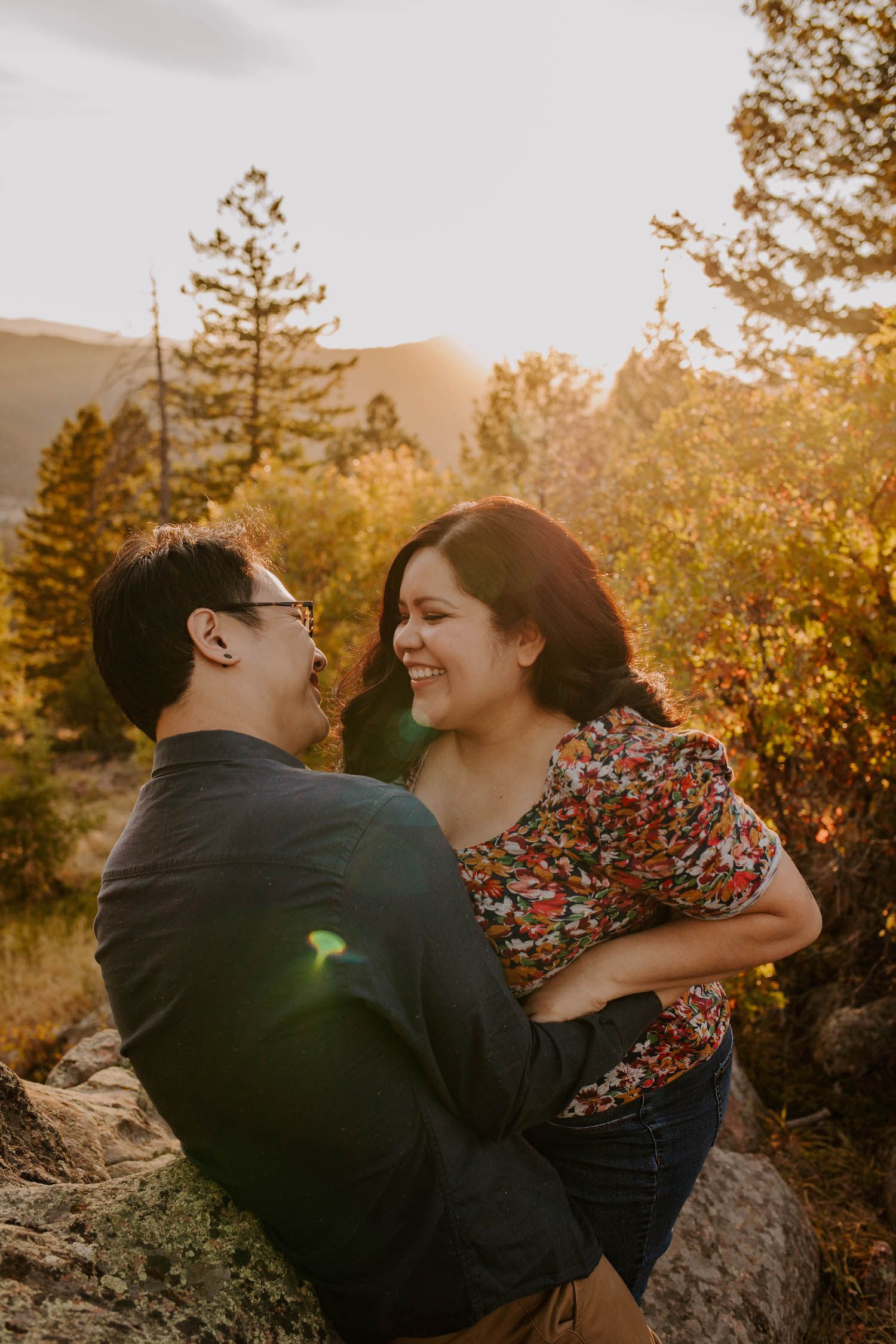 A couple shares a candid laugh together while sitting on rocks during golden hour in a mountain setting.