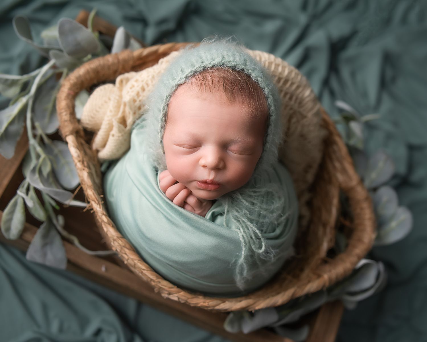 Newborn baby wrapped in mint green swaddle sleeping peacefully in a rustic wooden basket with soft floral decor.