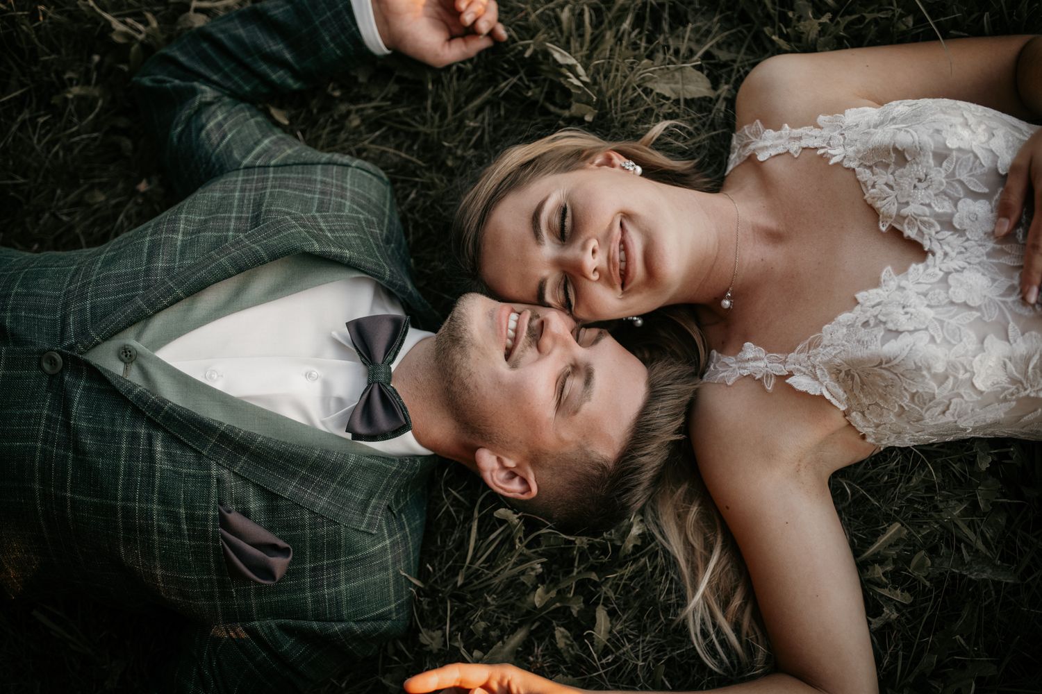 Aerial view of smiling bride and groom lying head to head on grass in wedding attire.