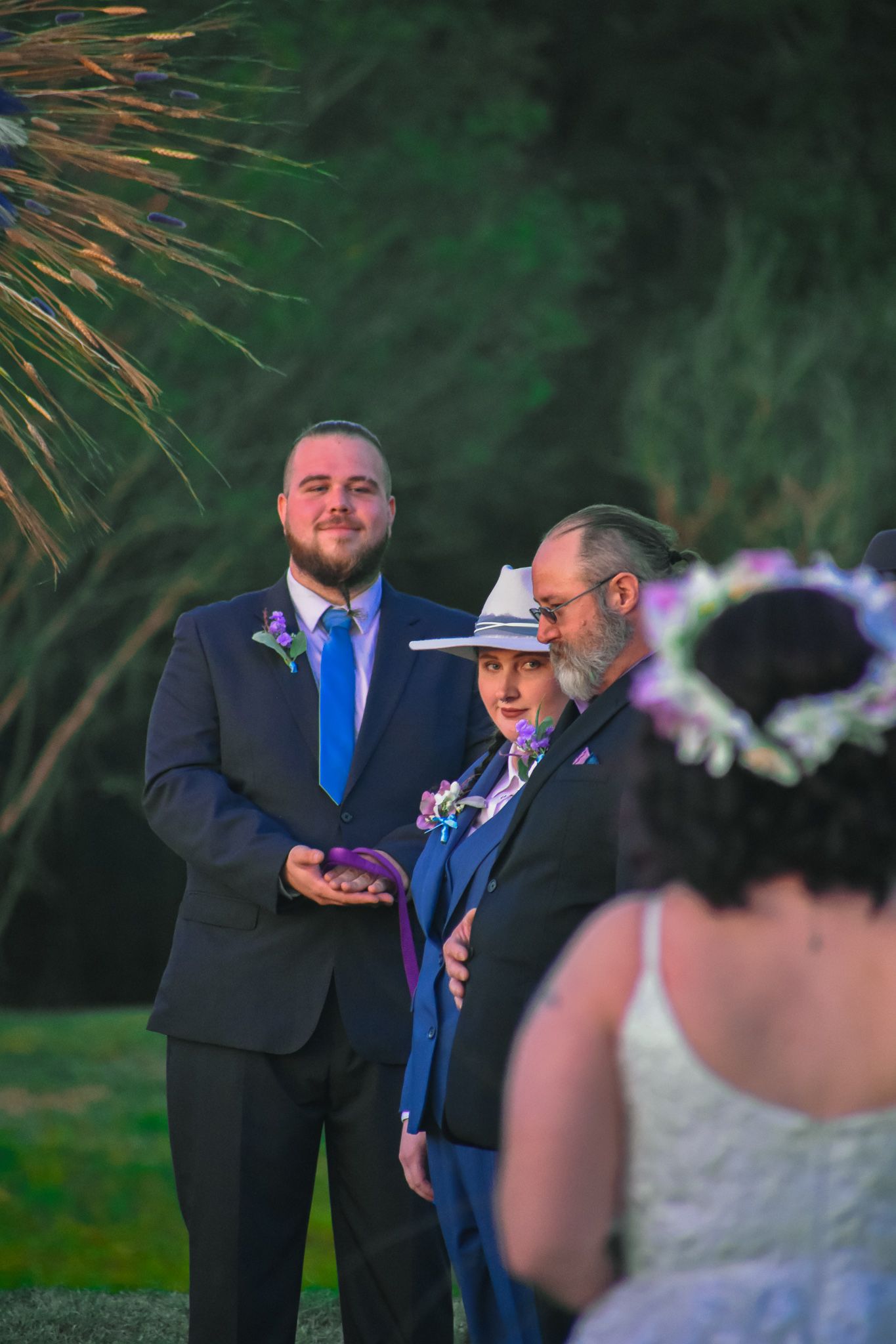 An outdoor wedding ceremony scene with attendees gathered under palm fronds at dusk.