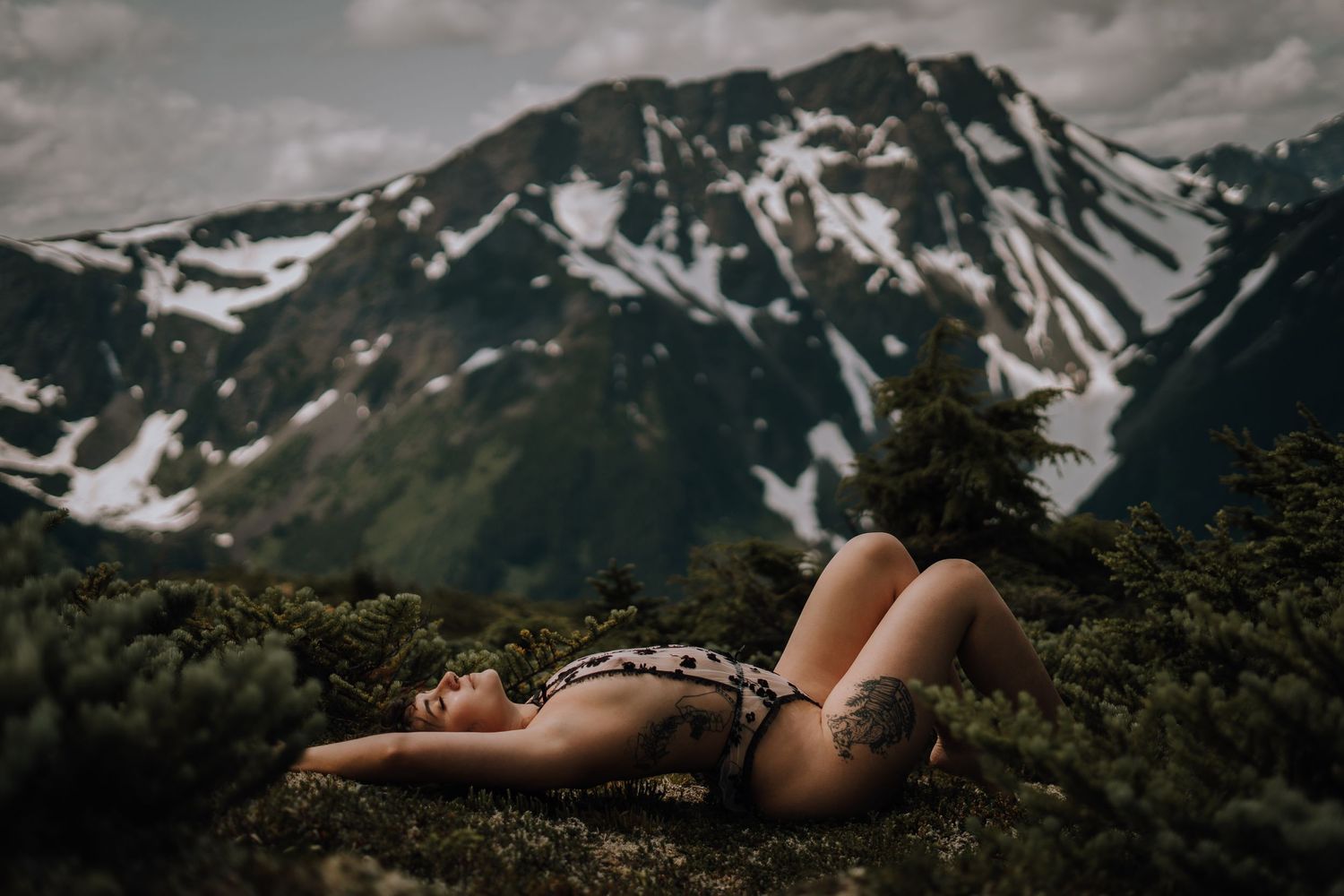 Artistic nude silhouette against dramatic mountain landscape with snow caps.
