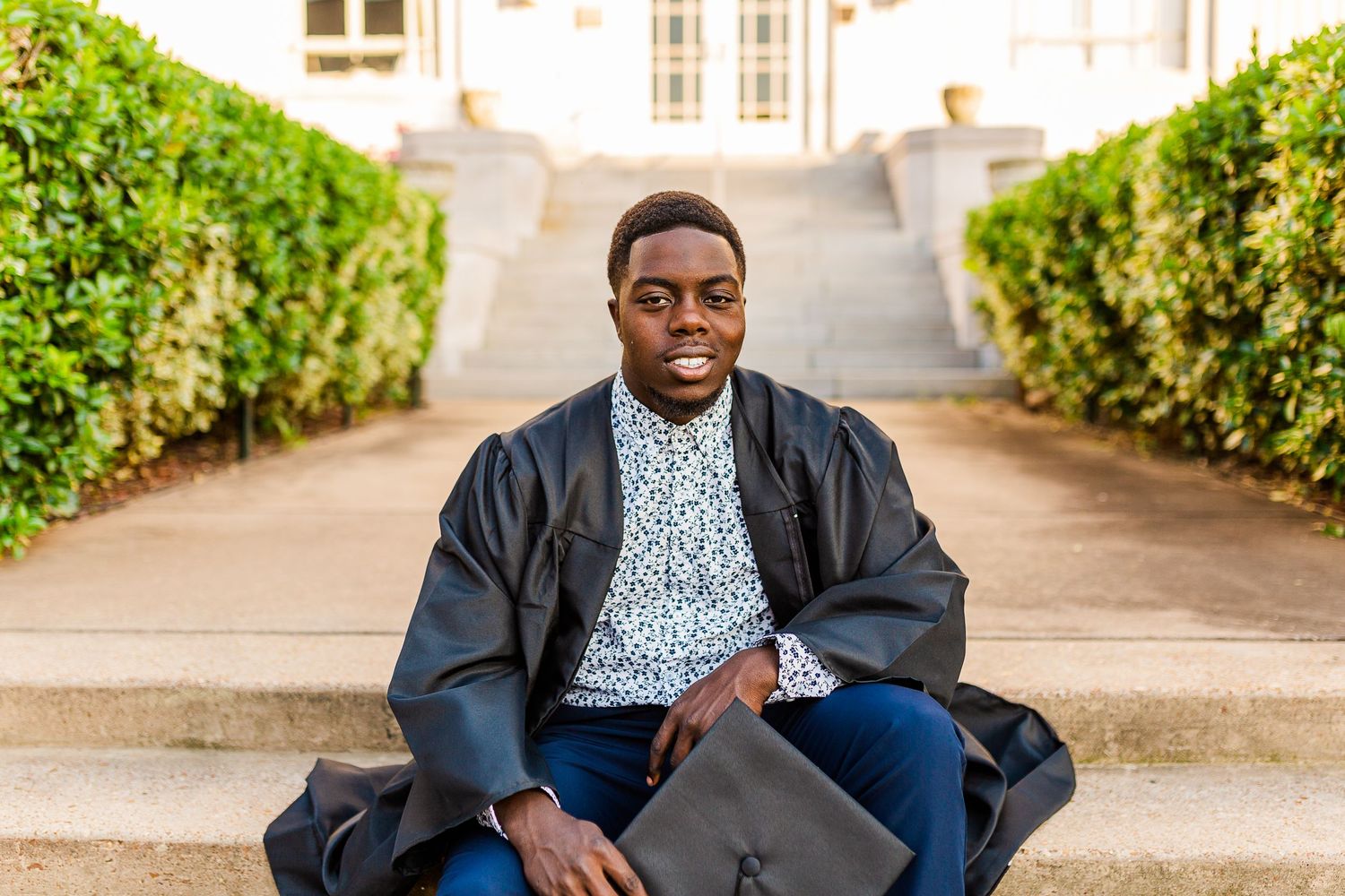 Student in graduation gown and glasses sits thoughtfully on steps between hedges.