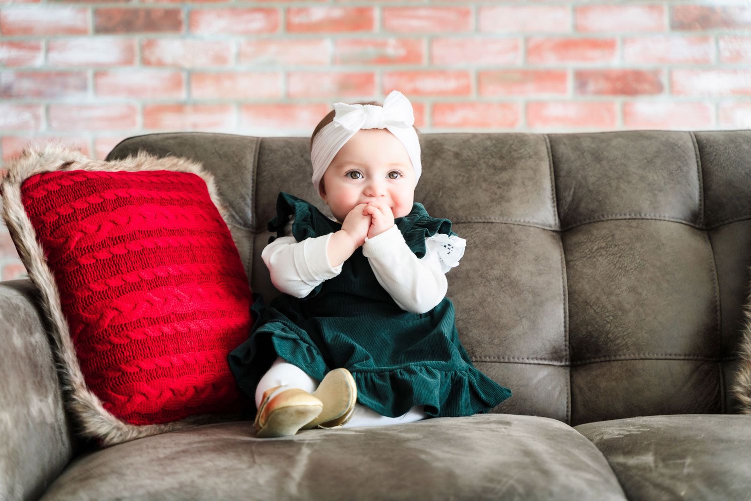 A baby in a green dress and white headband sits on a gray couch with red pillows.