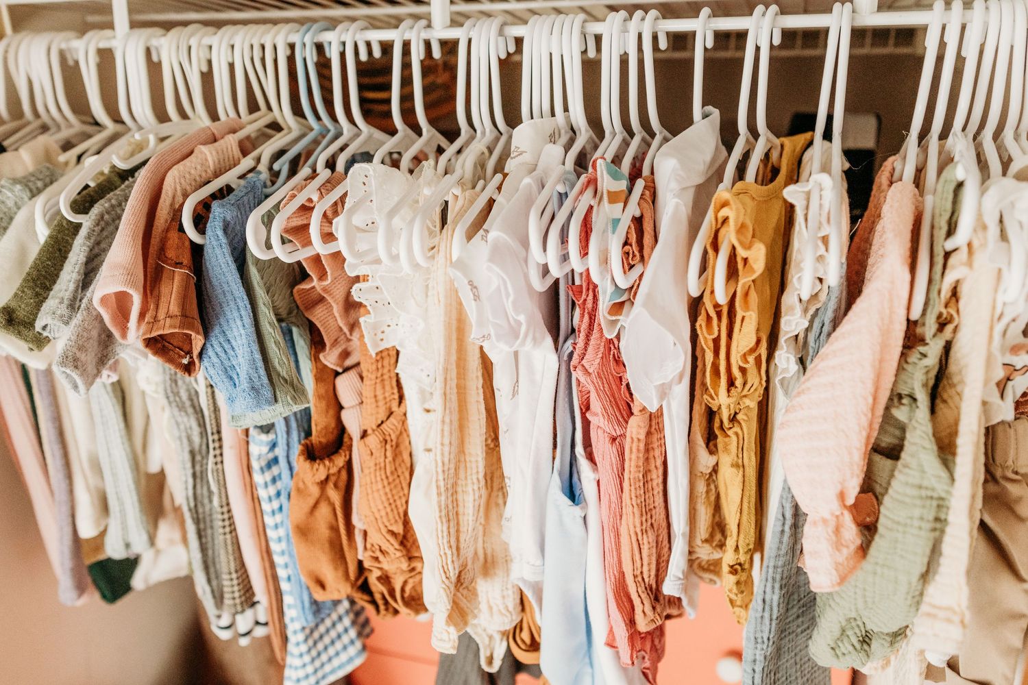 A closet rack filled with pastel and neutral-colored clothing on white hangers arranged neatly in a row.