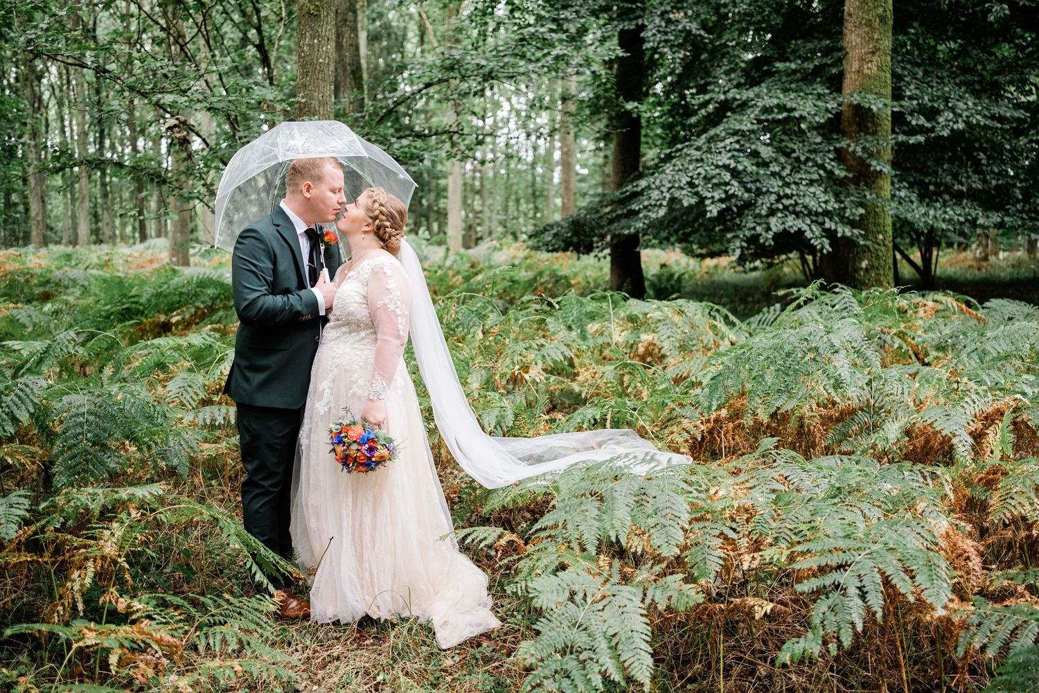 Newlyweds embrace under an umbrella in a lush forest setting with ferns.