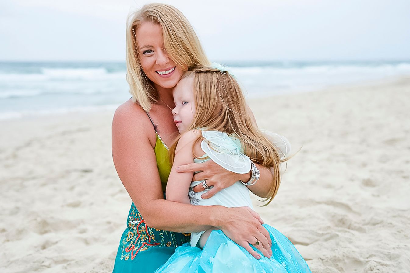 Two people share a joyful embrace on a sunny beach with ocean waves in the background.