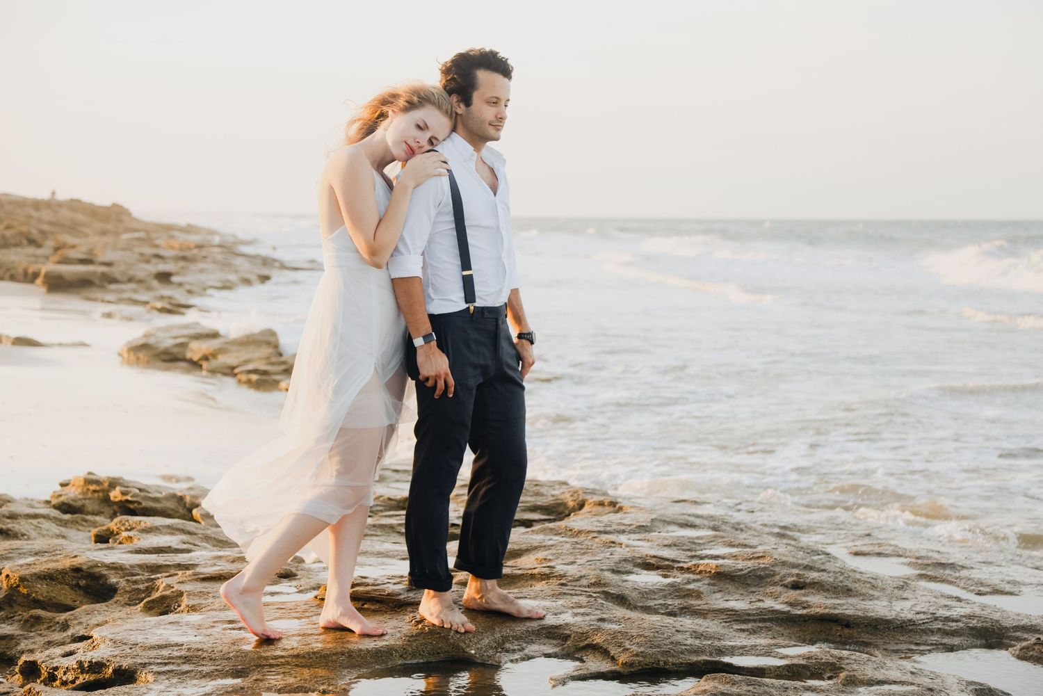 A couple stands together on rocky coastal shores at sunset.