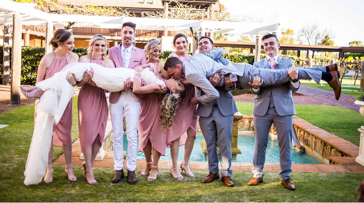 Wedding party in pastel pink and blue attire poses playfully on a lawn with a fountain.