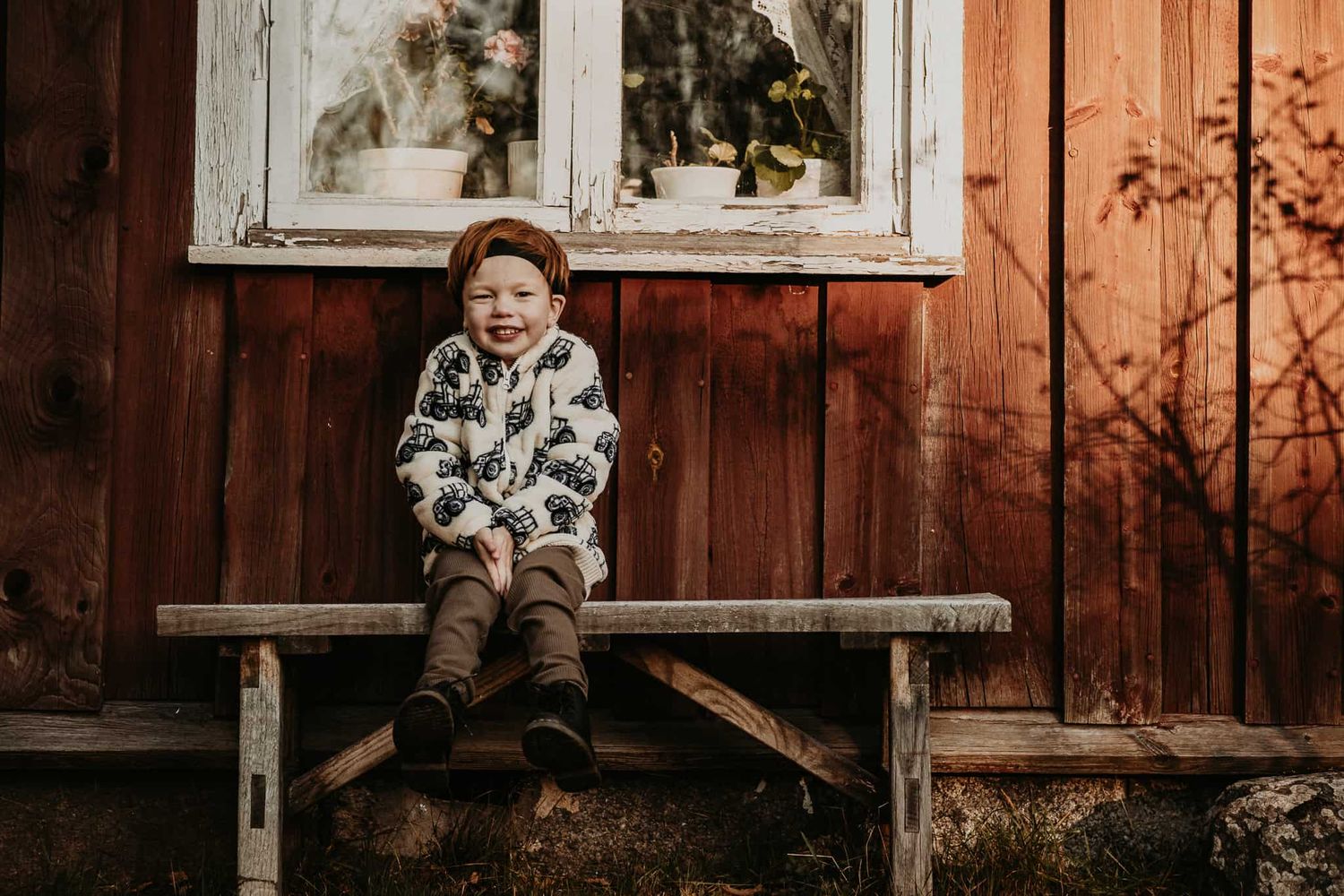 Pojke sitter och ler på en bänk utanför ett gammalt hus under en familjefotografering i Jönköping Mullsjö.