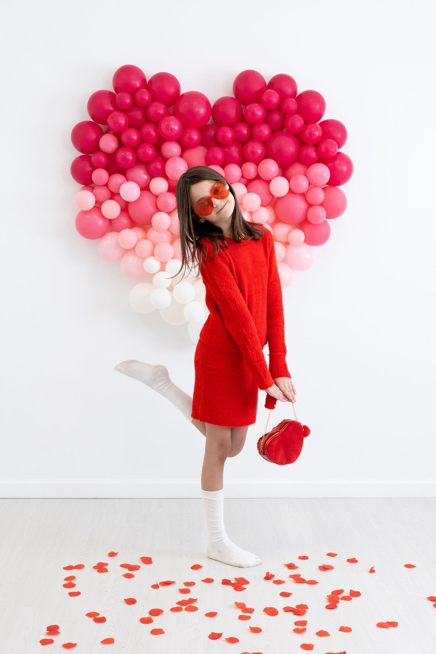 Festive Valentine's scene with red dress and pink ombre balloon heart backdrop on white wall with scattered petals.