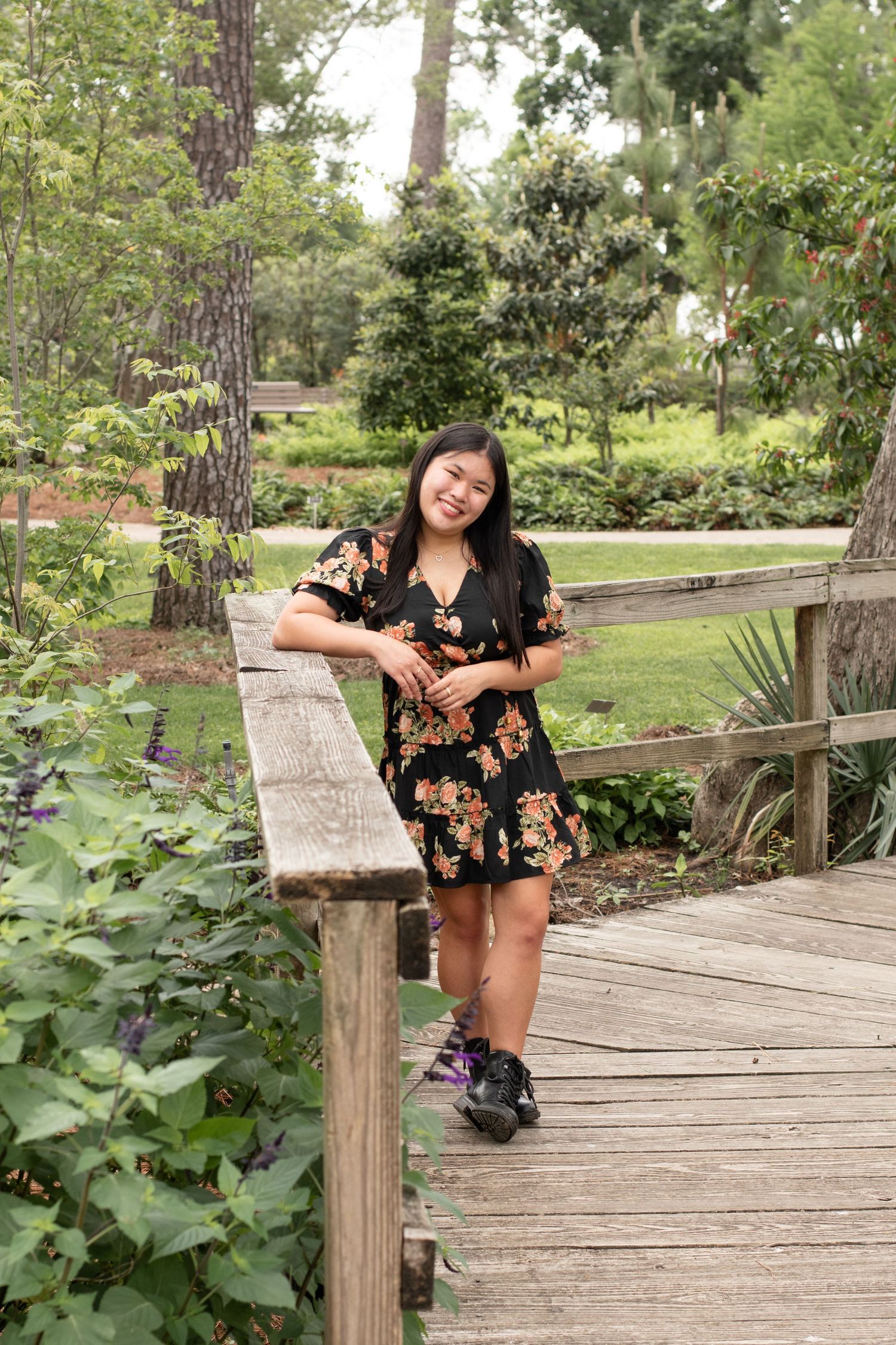 A woman in a floral dress poses casually on a wooden bridge railing in a lush garden setting.