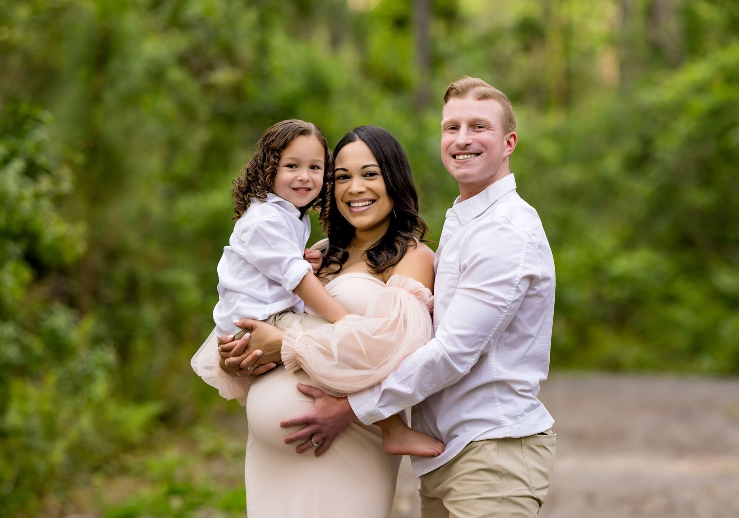 Family poses together outdoors in white and beige outfits against lush green foliage.