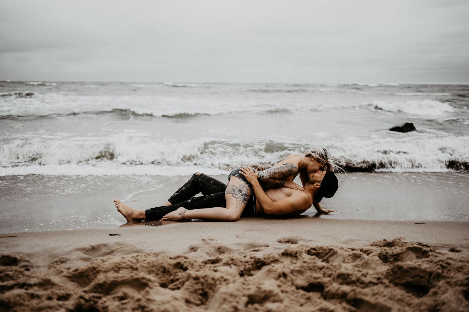 Couple embracing on sandy beach with ocean waves in moody overcast weather.
