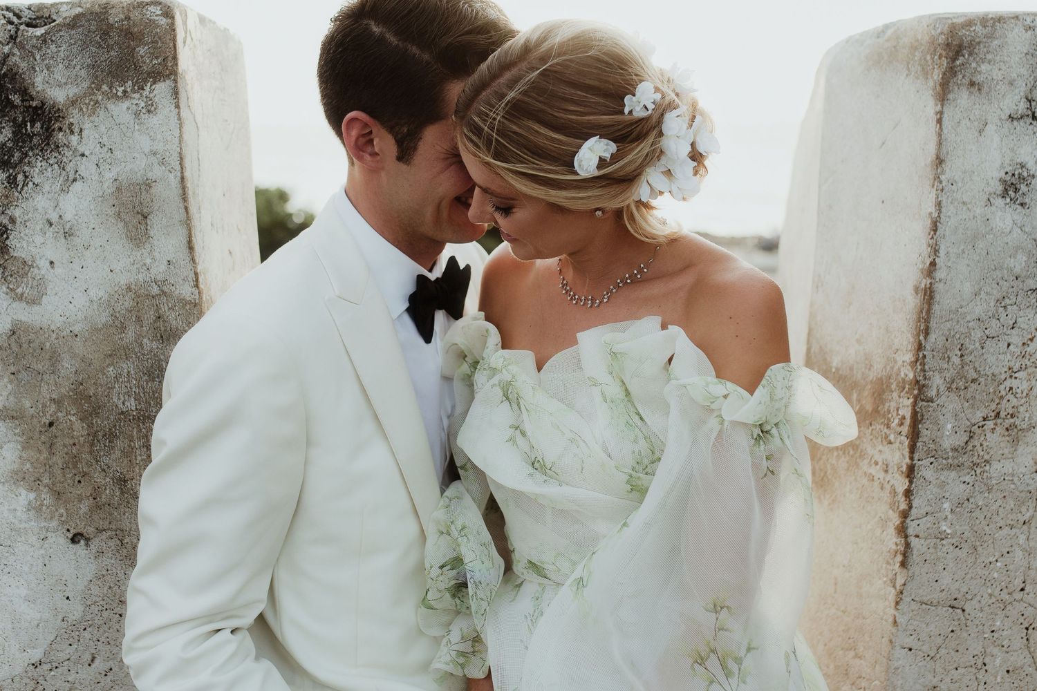 Intimate wedding moment captured against white stone wall with delicate bridal details.
