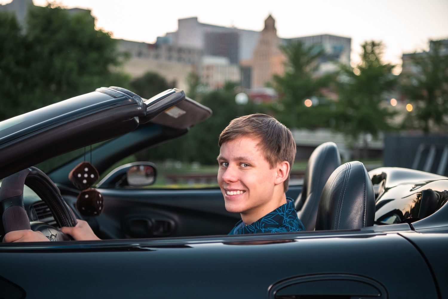 Portrait of someone smiling while sitting in a black convertible car at dusk.