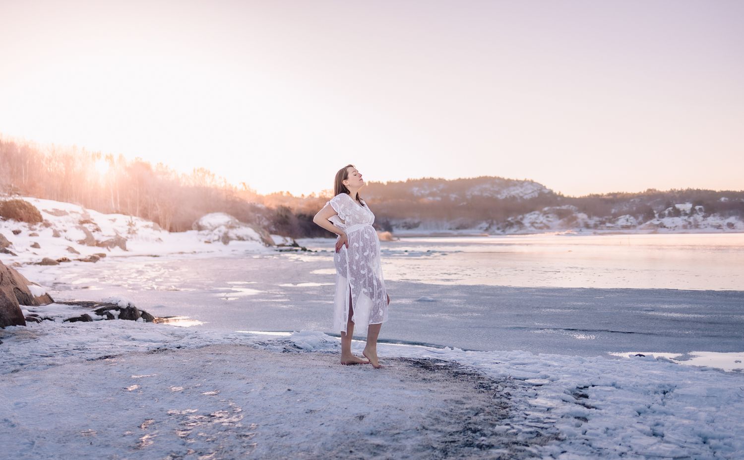 Figure in white stands on snowy beach at sunset with mountains in background.