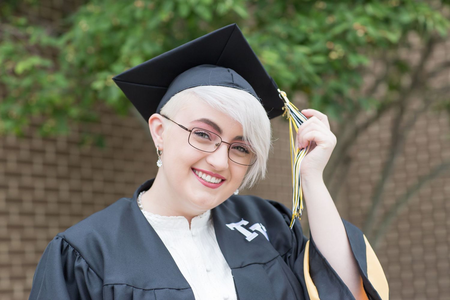 Smiling graduate in black cap and gown with blonde hair and glasses holding tassel outdoors.