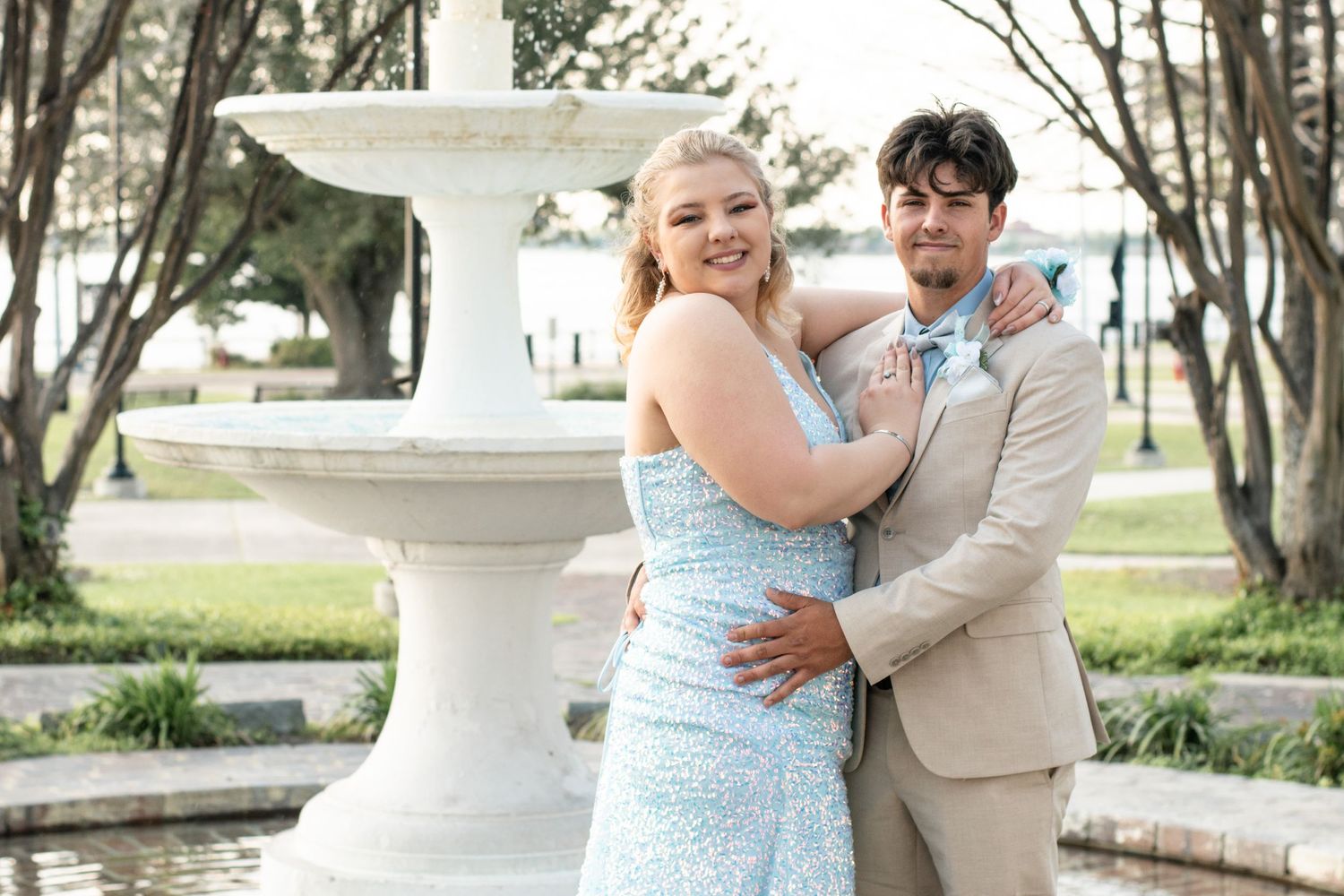 A couple in matching formal attire poses by a white fountain in a garden setting.