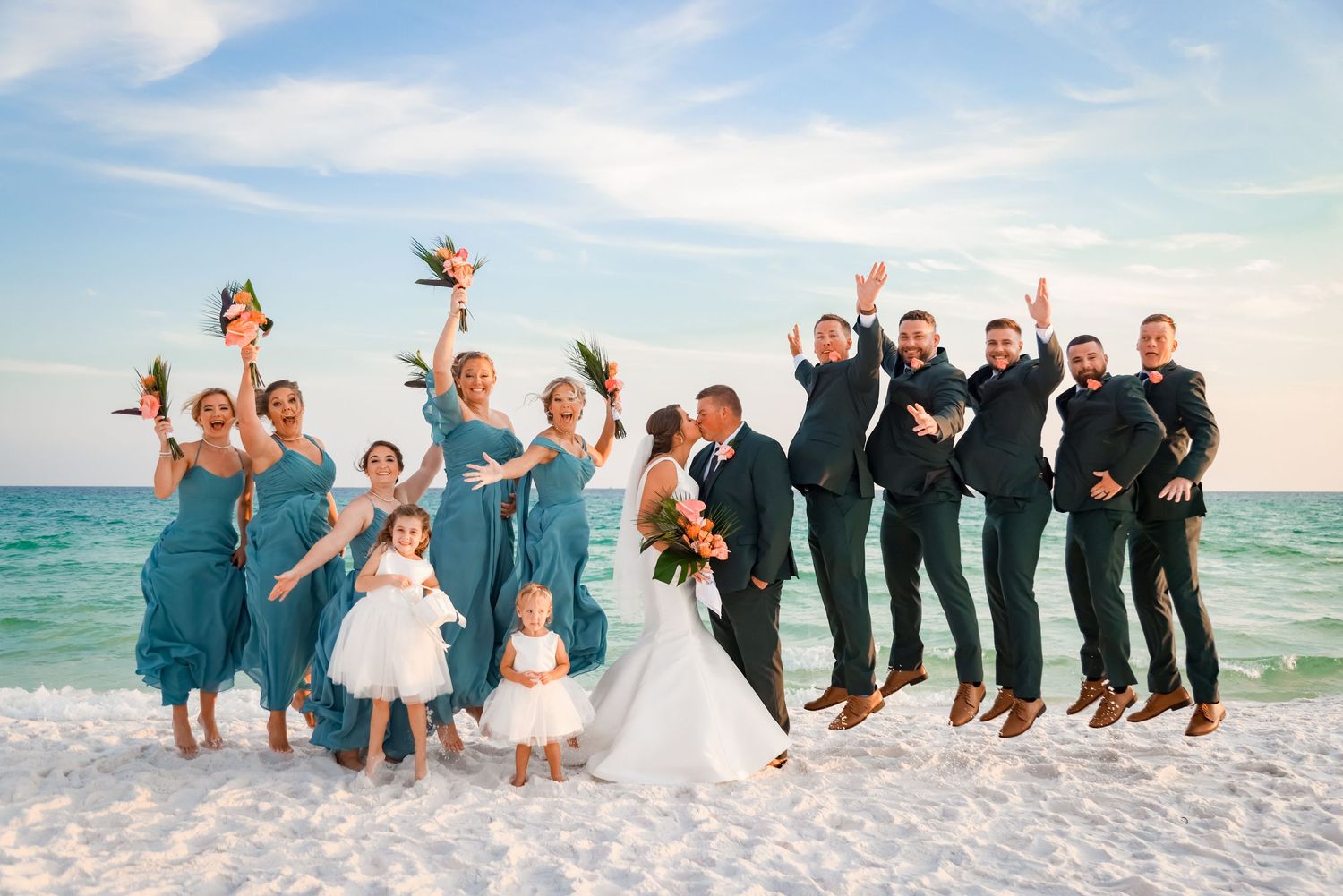 Joyful wedding party jumping on beach with ocean backdrop.
