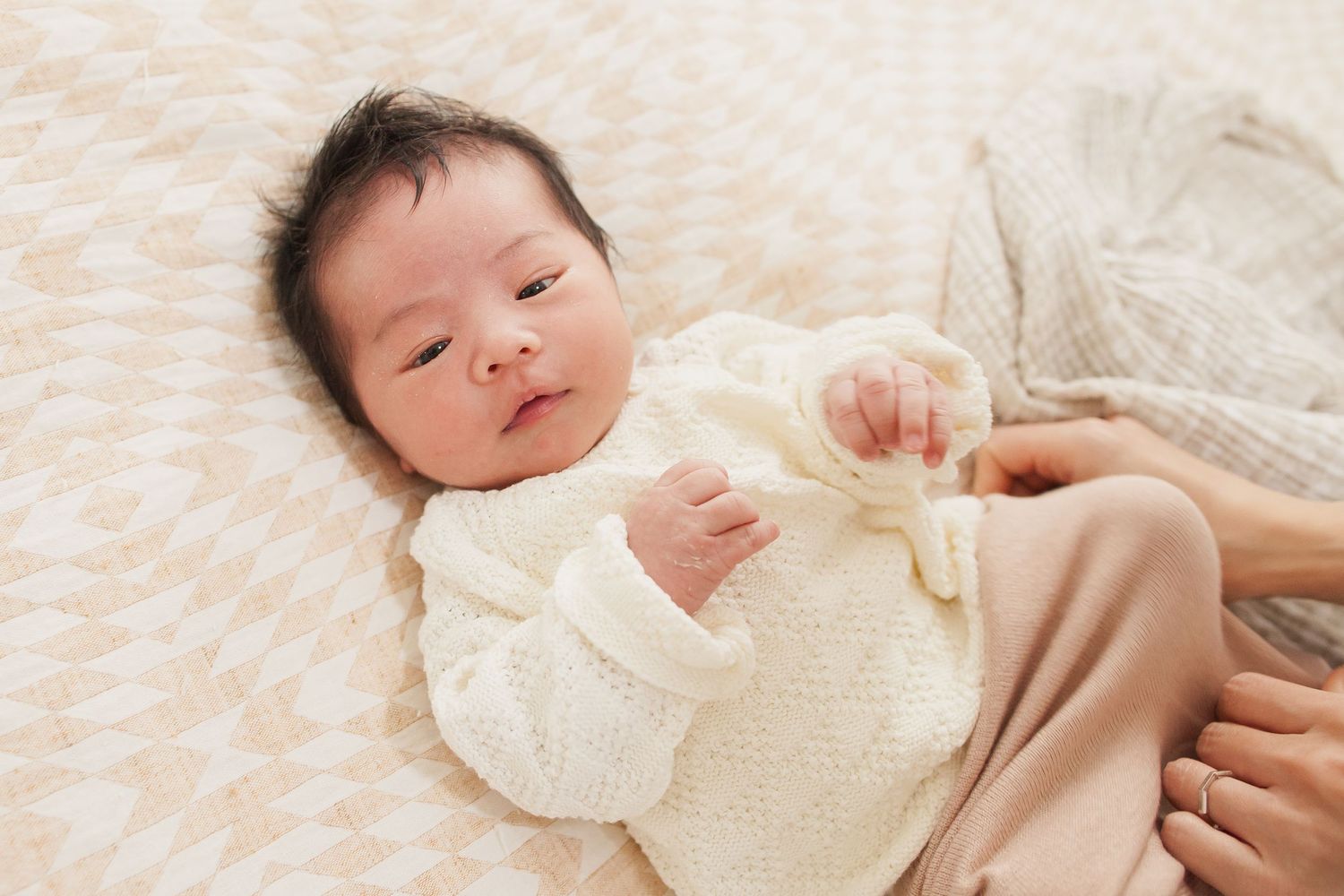 Newborn baby wearing a white sweater lying on a textured blanket.