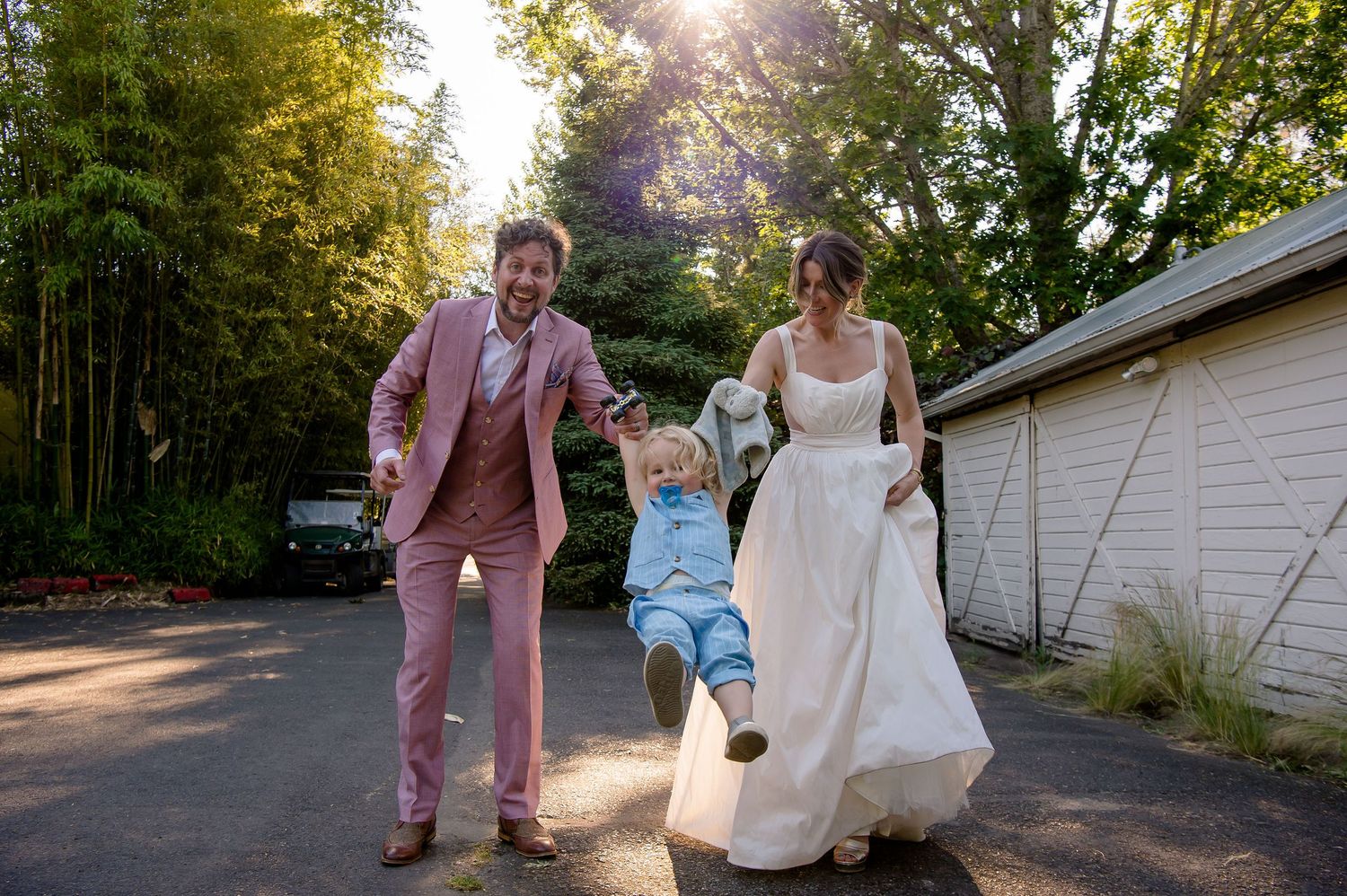 Wedding couple swings a child between them on a sunny outdoor path.