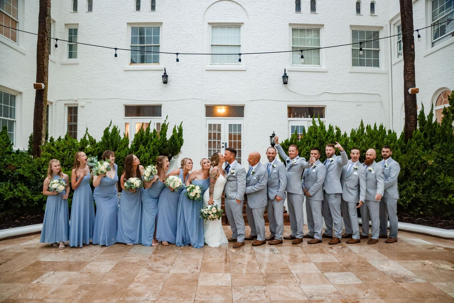 Wedding party in coordinating blue dresses and grey suits poses against white building exterior.