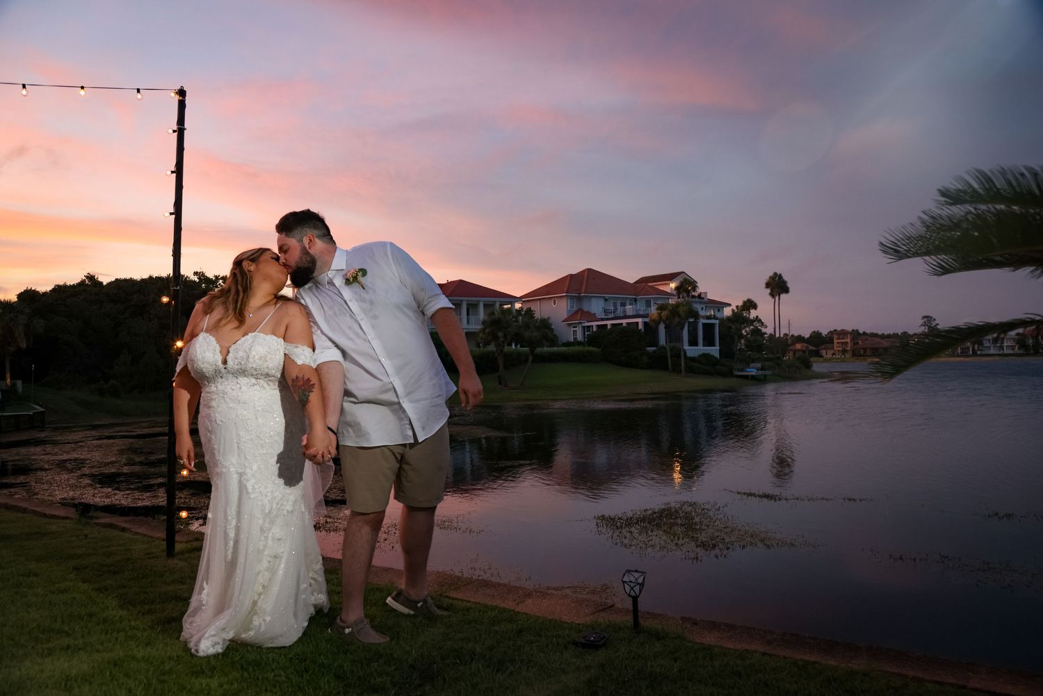 A romantic sunset moment by a lakeside with string lights and houses in the background.