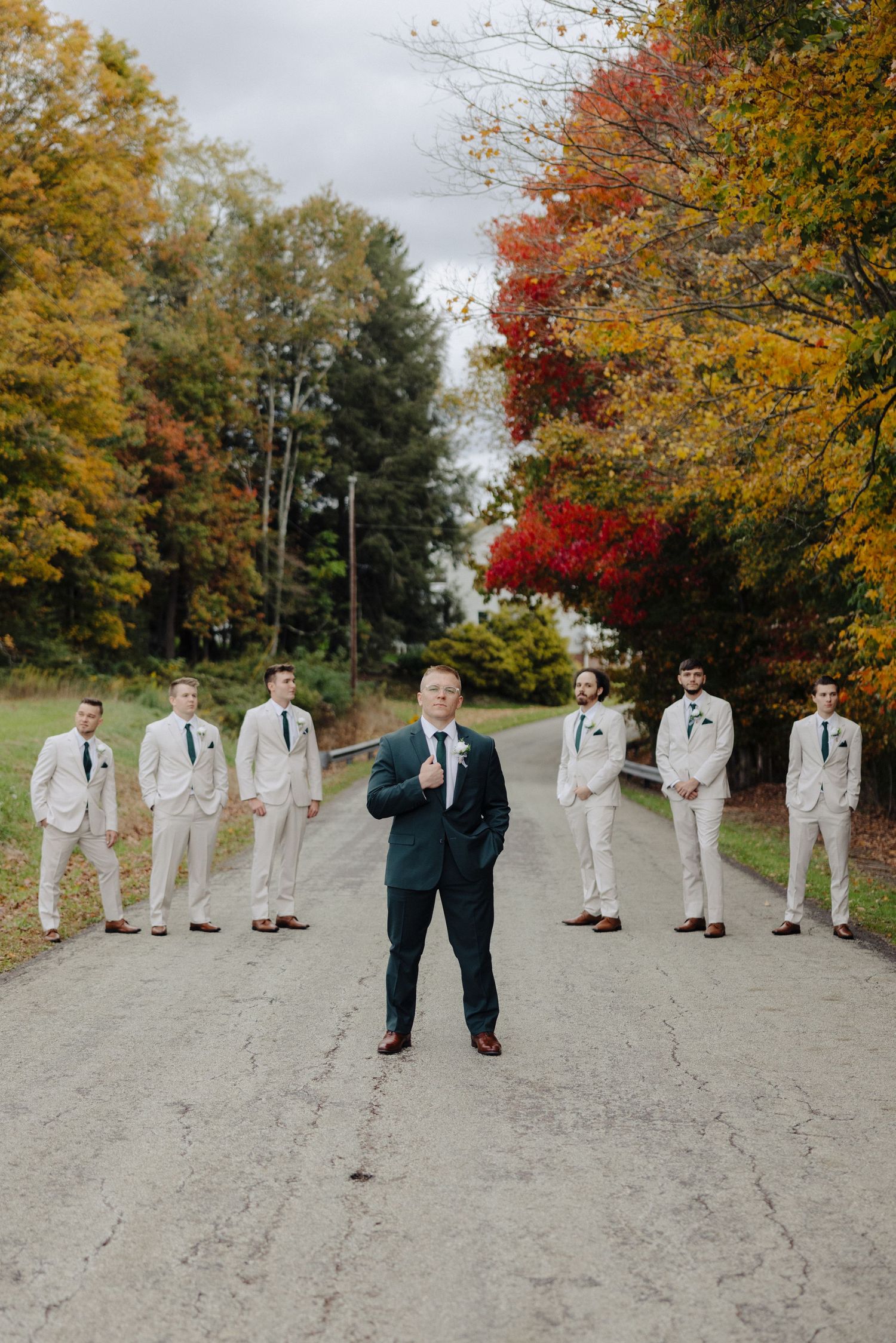 Groomsmen in light suits stand behind groom in navy on path surrounded by vibrant autumn foliage.