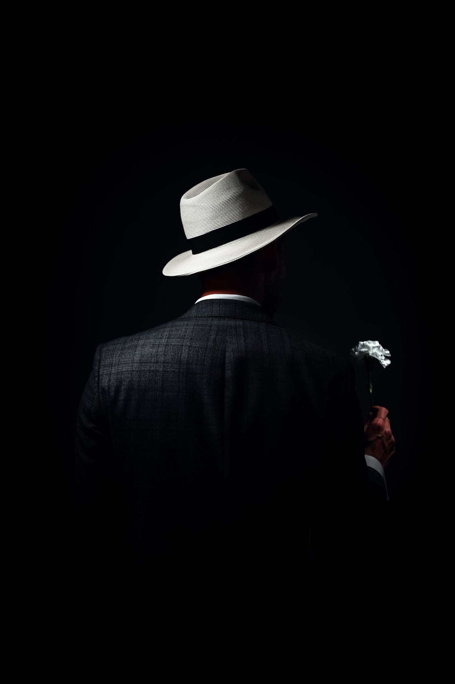 Man in white fedora hat and dark suit holding a cigar, viewed from behind against black background.