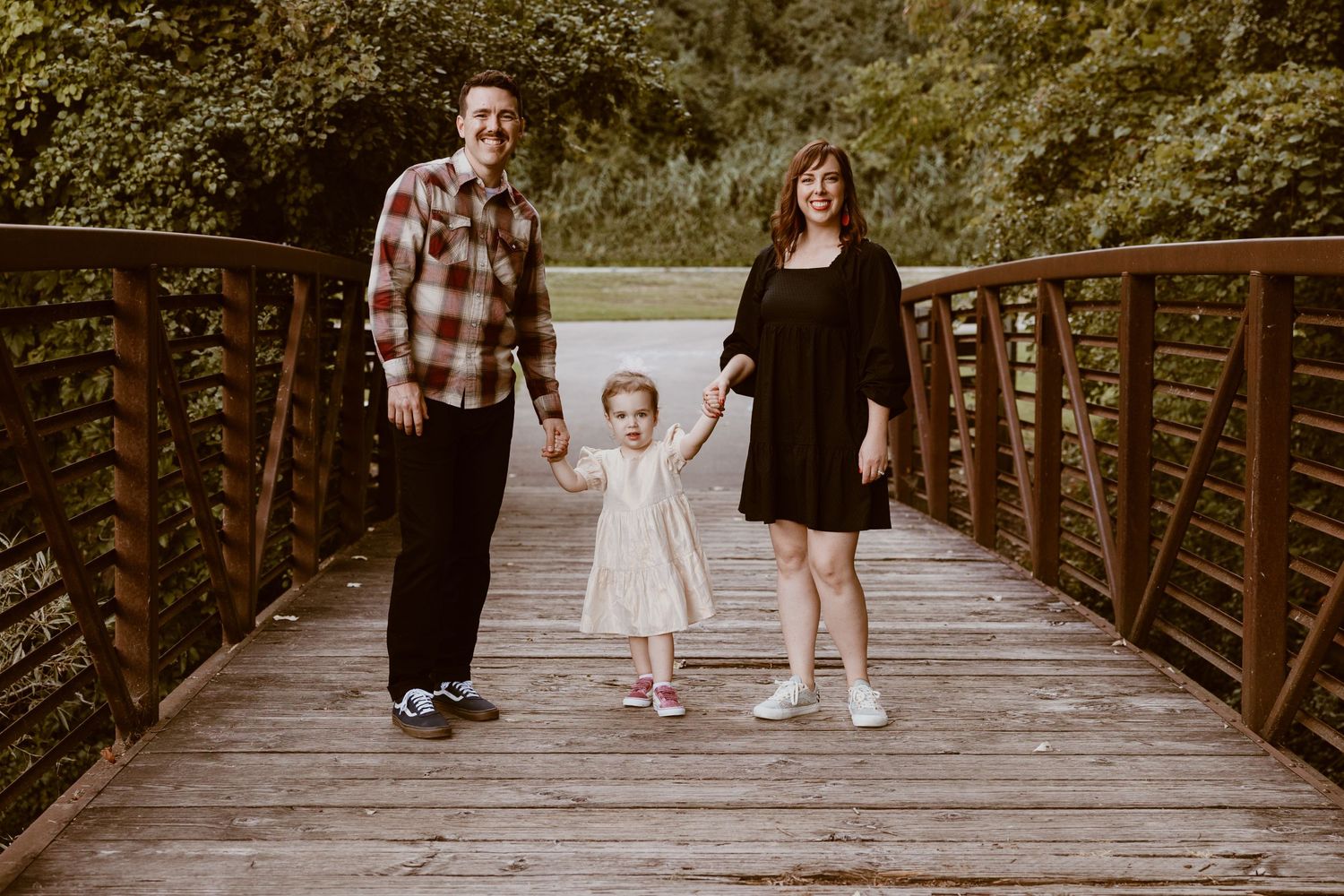 A family poses together holding hands on a wooden bridge in a park setting.