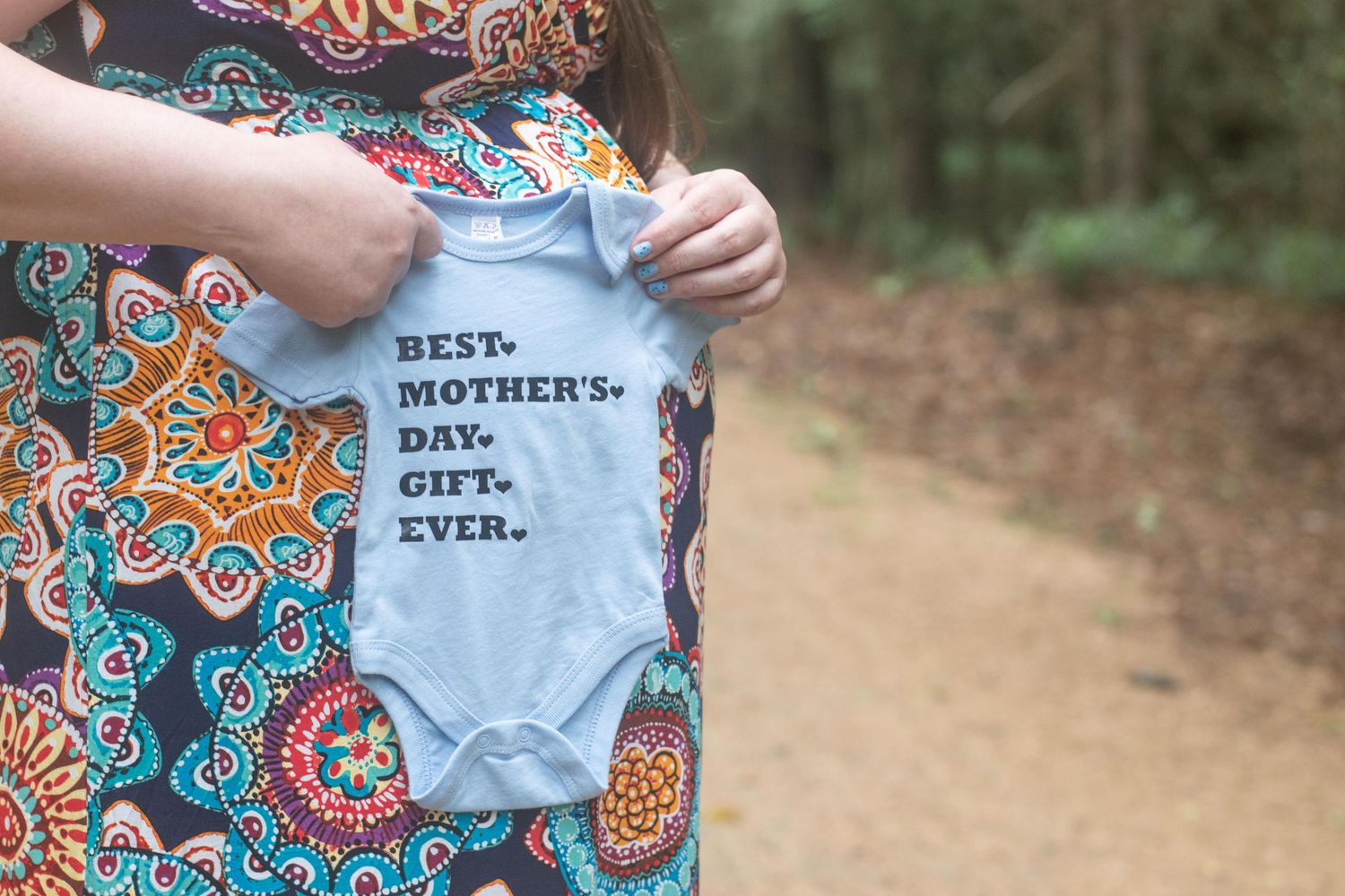 Hands holding up blue baby onesie with text against colorful patterned dress background.