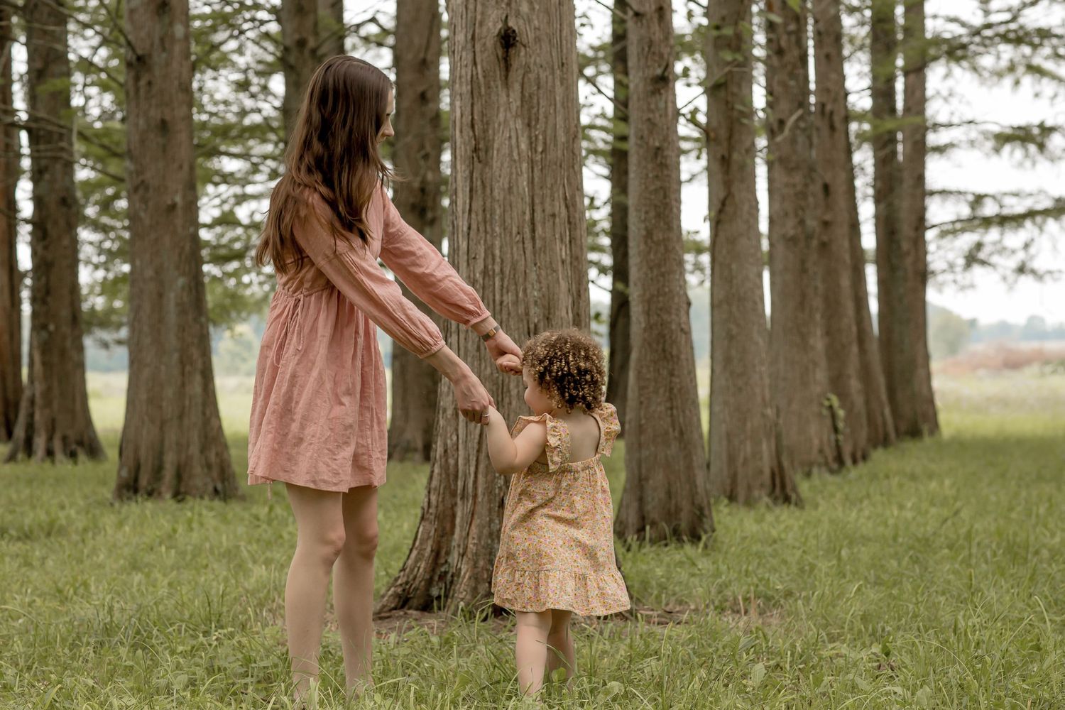 A mother and young daughter hold hands while walking among tall trees in a lush green park.