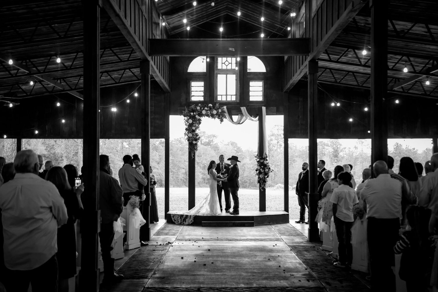 Silhouetted figures stand in a rustic barn doorway surrounded by wedding guests during a ceremony.