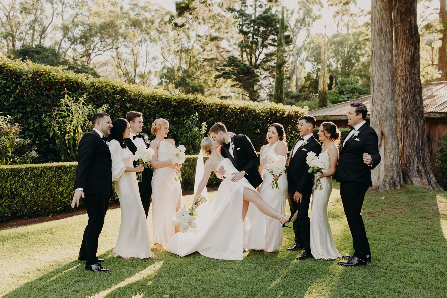 Wedding party relaxing on grass under trees during an outdoor celebration.