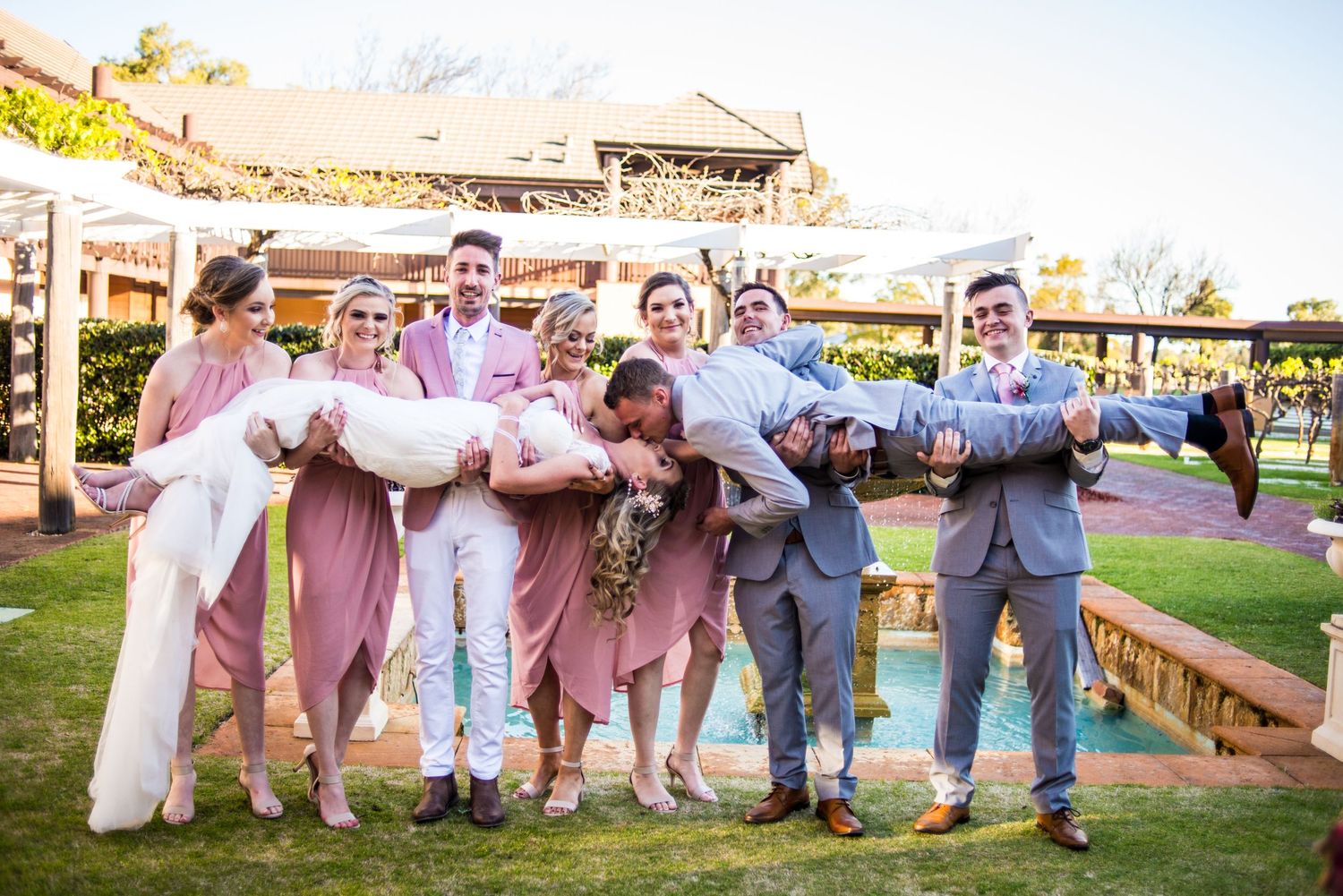 Wedding party in pink and gray attire posing playfully in an outdoor garden setting.